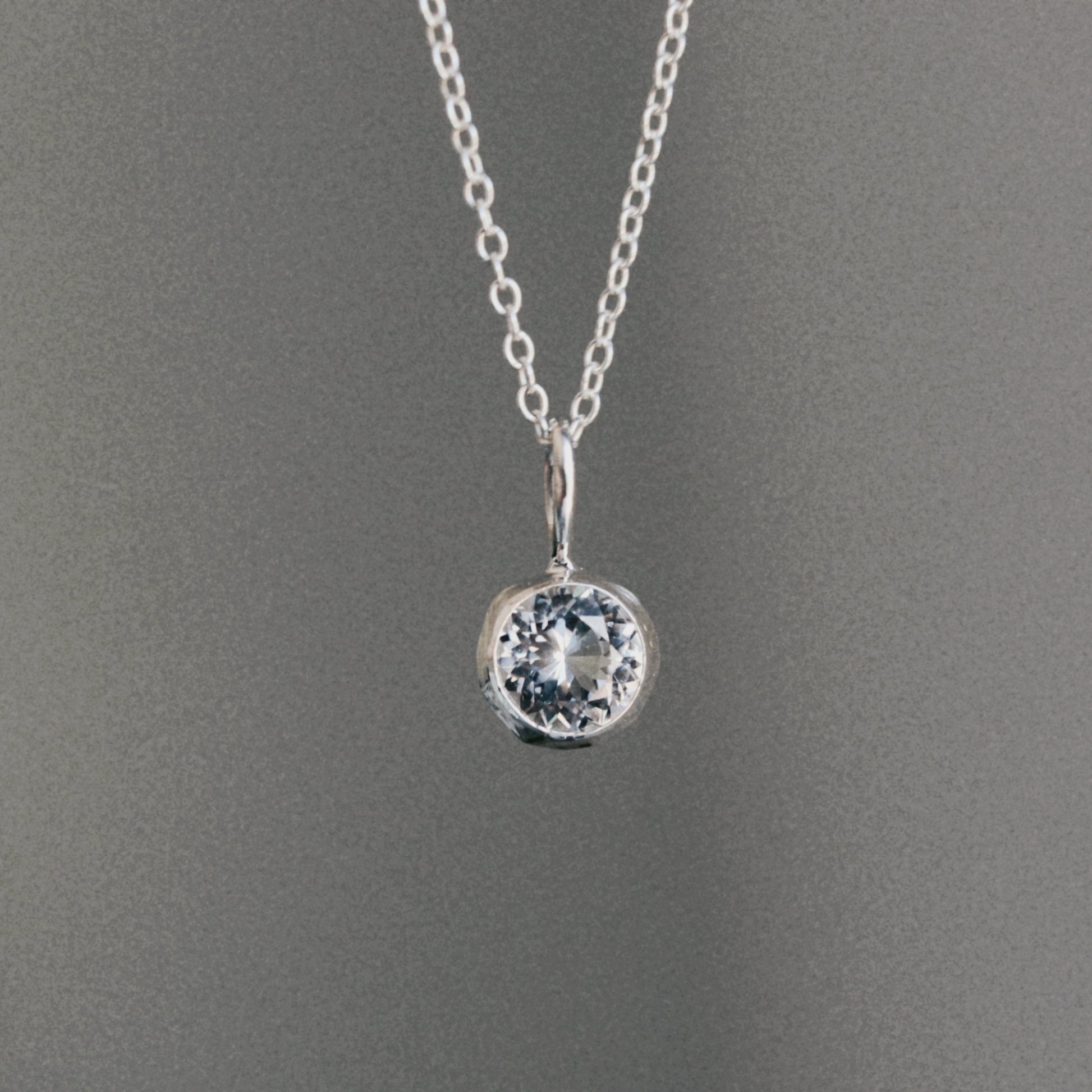 A silver chain necklace with a round bezel-set clear gemstone pendant, displayed against a neutral gray background.