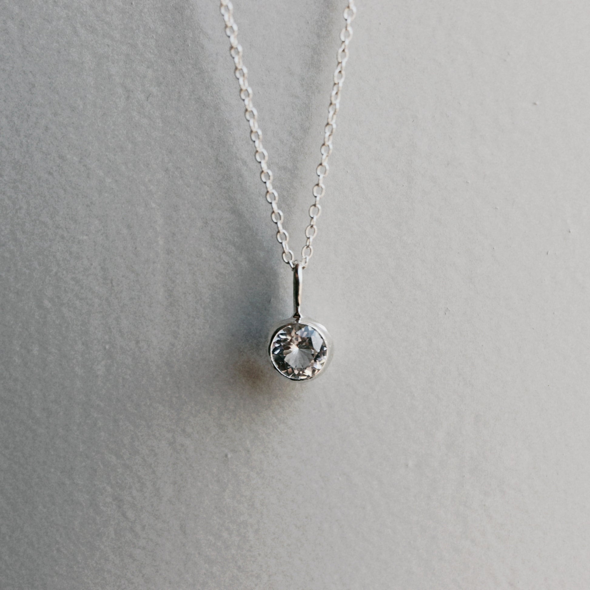 A delicate silver chain necklace with a single bezel-set clear round gemstone pendant, displayed against a neutral light gray textured background.