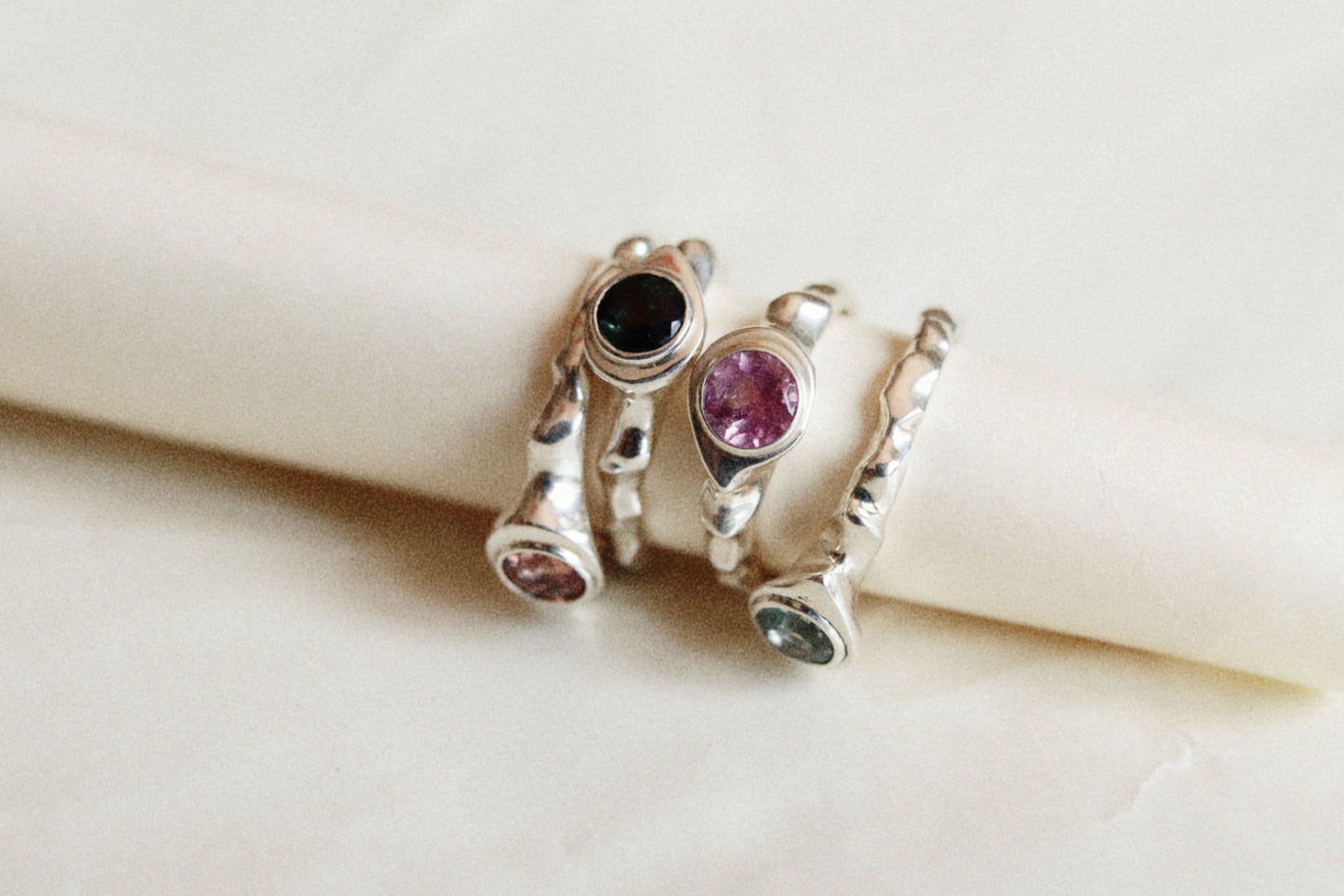 Three silver rings with colorful gemstones displayed on a cream-colored ring holder. The rings feature round bezel-set stones in black, pink, and green.