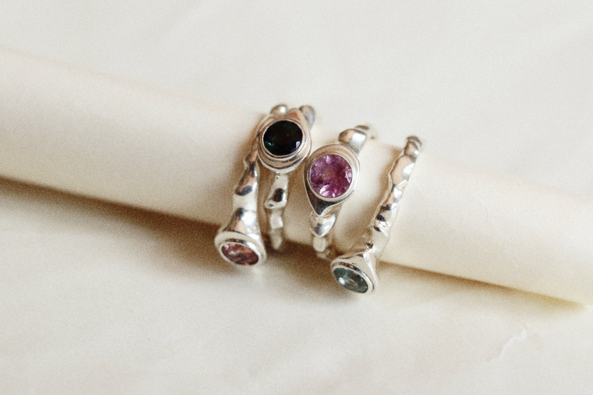 Three silver rings with colorful gemstones displayed on a cream-colored ring holder. The rings feature round bezel-set stones in black, pink, and green.