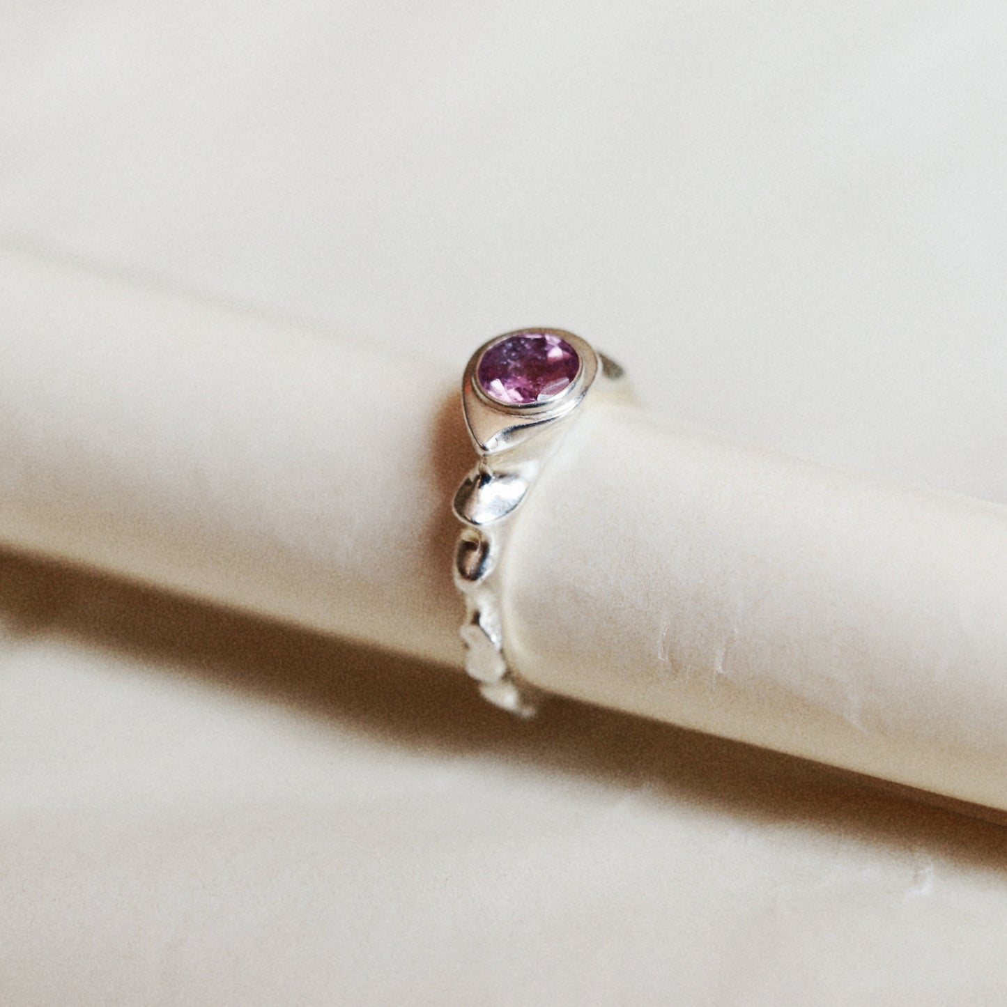 A silver ring with a round pink gemstone, displayed on a cream-colored ring stand against a soft beige background.