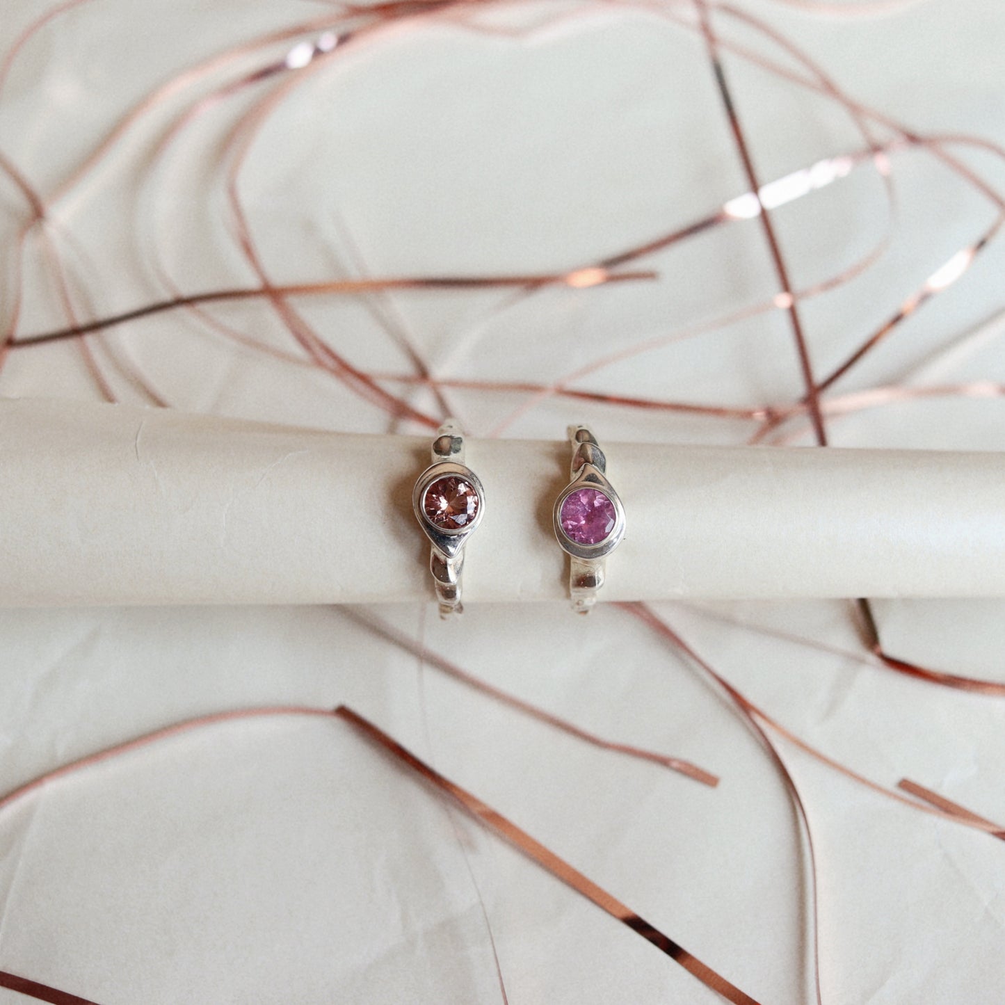 Two silver rings with round pink gemstones, displayed on a white cylindrical stand with rose gold wire accents in the background.