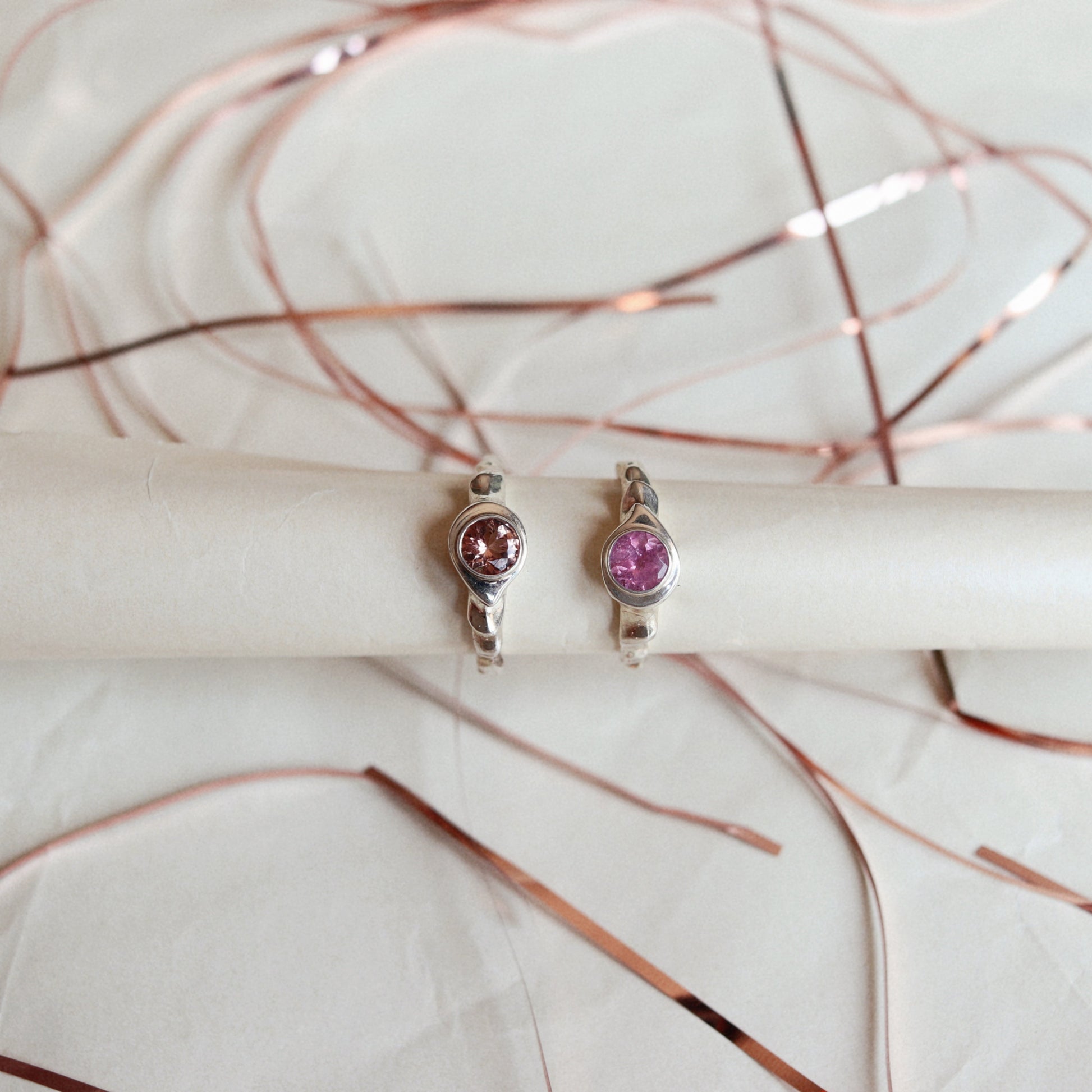 Two silver rings with round pink gemstones, displayed on a white cylindrical stand with rose gold wire accents in the background.
