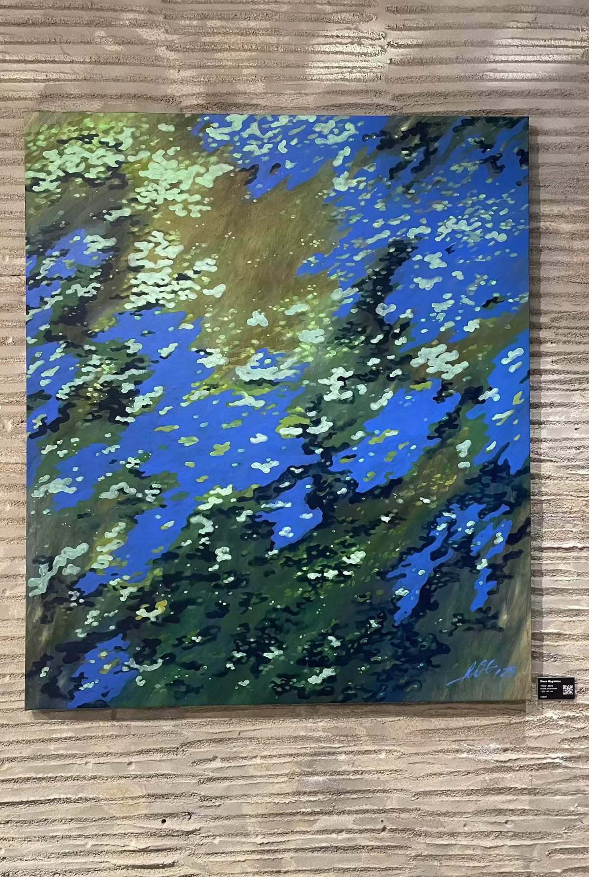 An abstract landscape painting on a square canvas, featuring vibrant blue, green, and brown brushstrokes, displayed on a textured wooden wall.