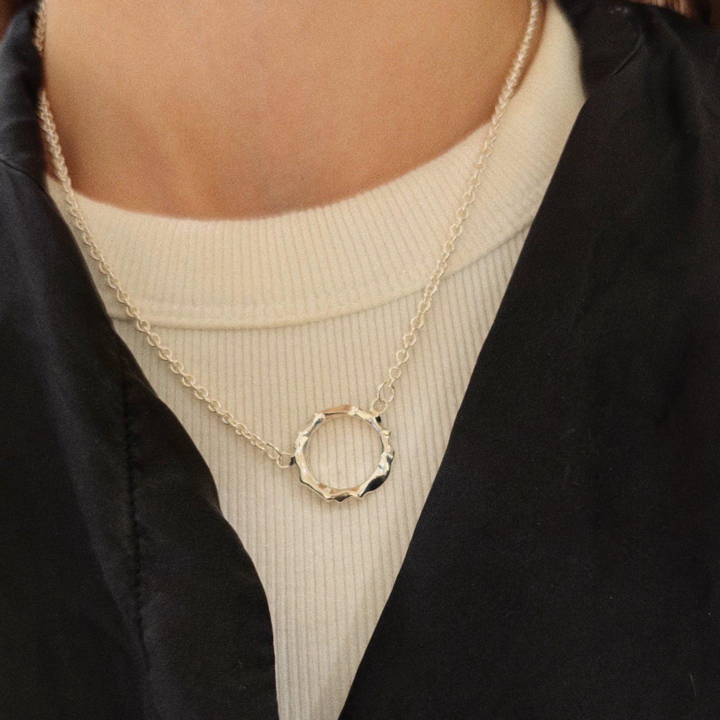 A close-up of a person wearing a delicate silver chain necklace with a faceted, textured circular pendant over a cream ribbed turtleneck and a black satin jacket.