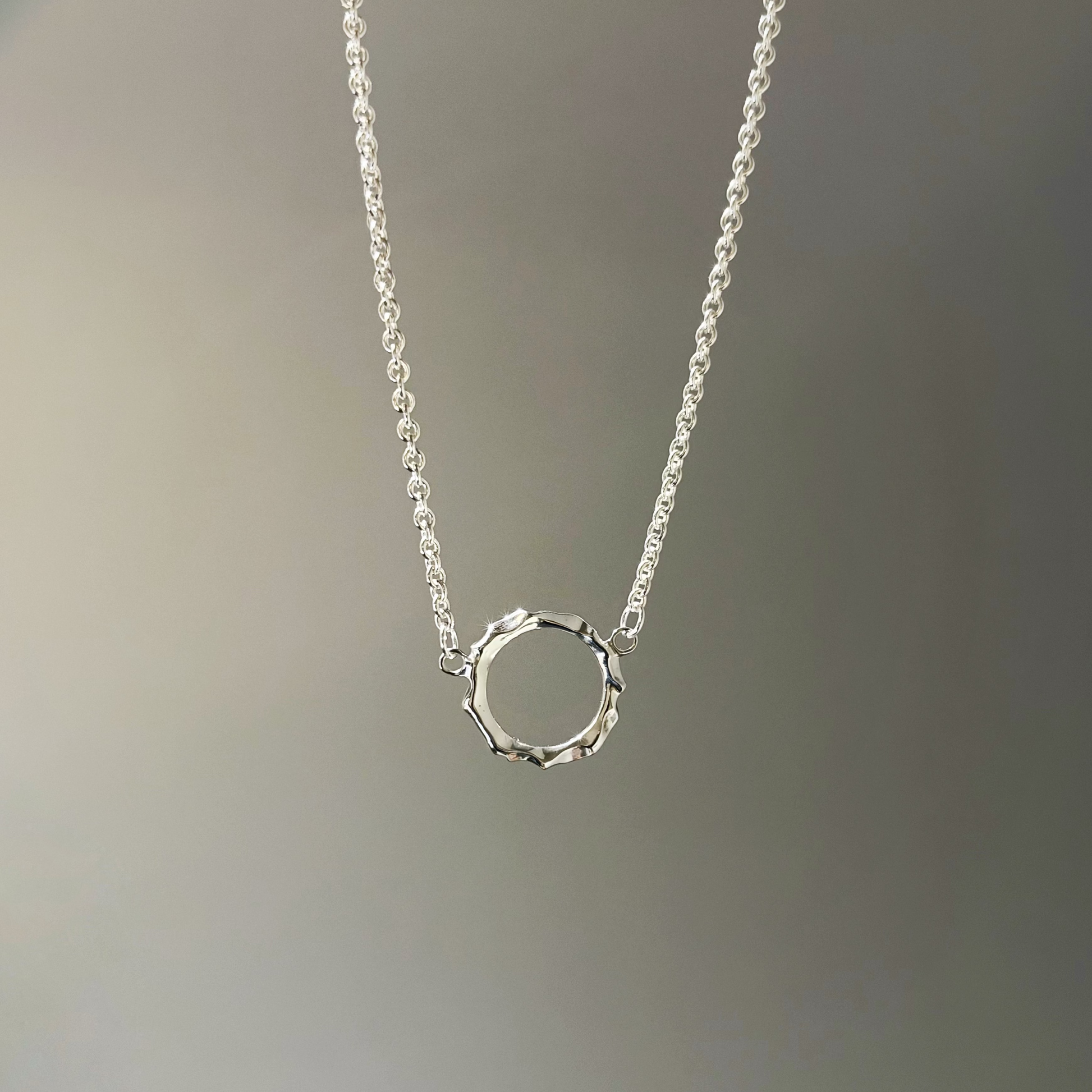 A delicate silver chain necklace with a small, textured open ring pendant, shown against a soft gray gradient background.