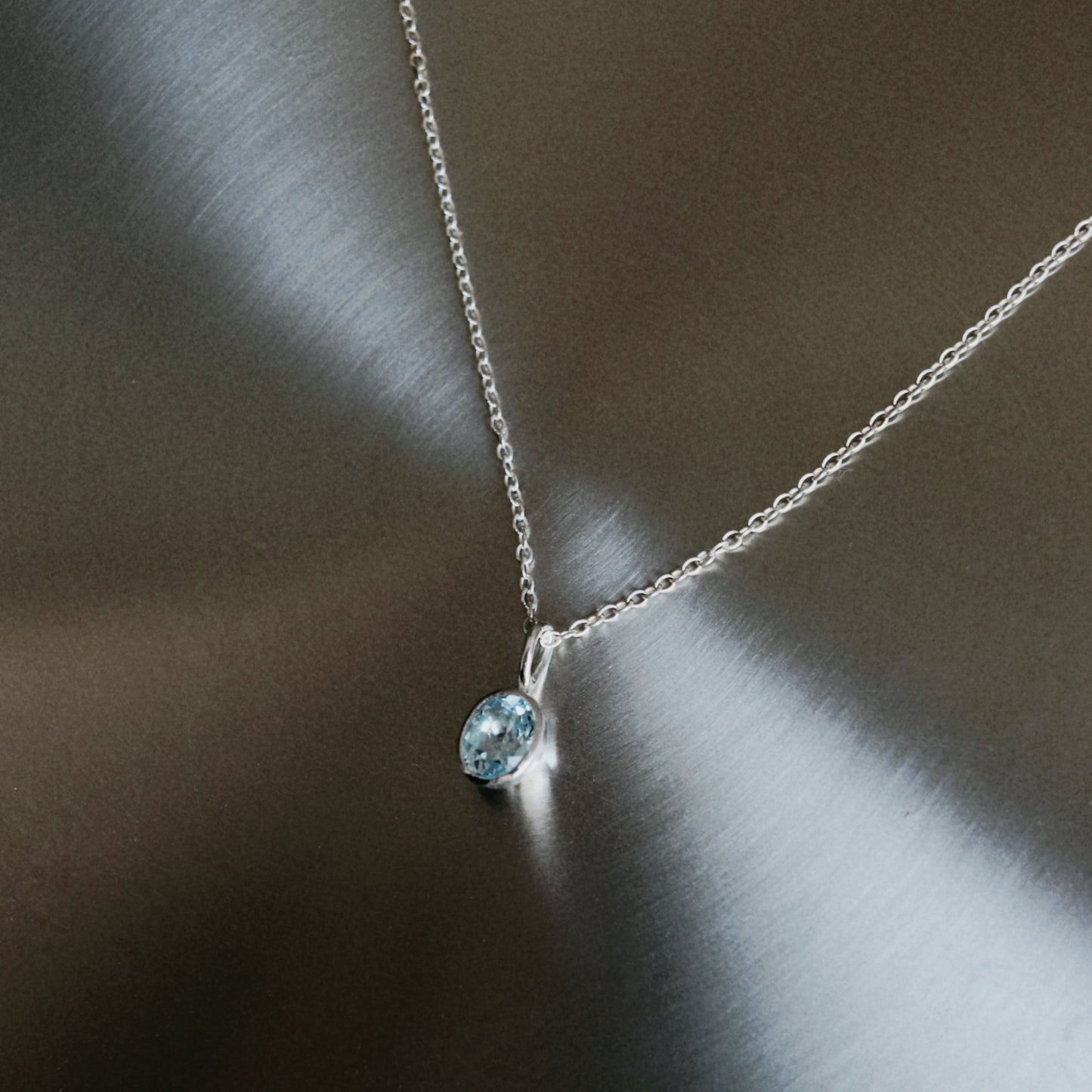 A delicate silver chain necklace with a small oval light blue gemstone pendant, displayed on a brushed metallic surface.