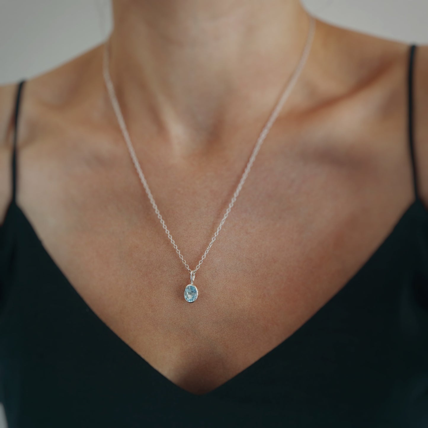 A close-up of a delicate silver chain necklace with a small oval light blue gemstone pendant, worn by a model in a black top.
