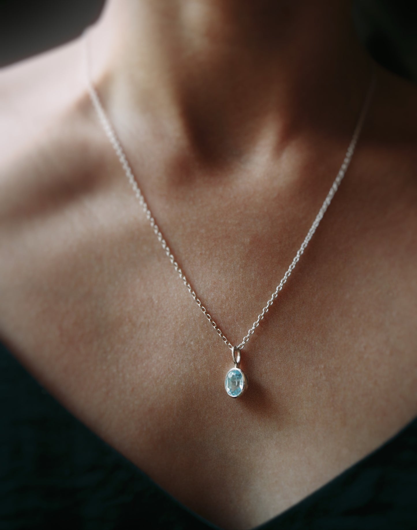 A delicate silver chain necklace with a small oval light blue gemstone pendant, worn on a model's neck.