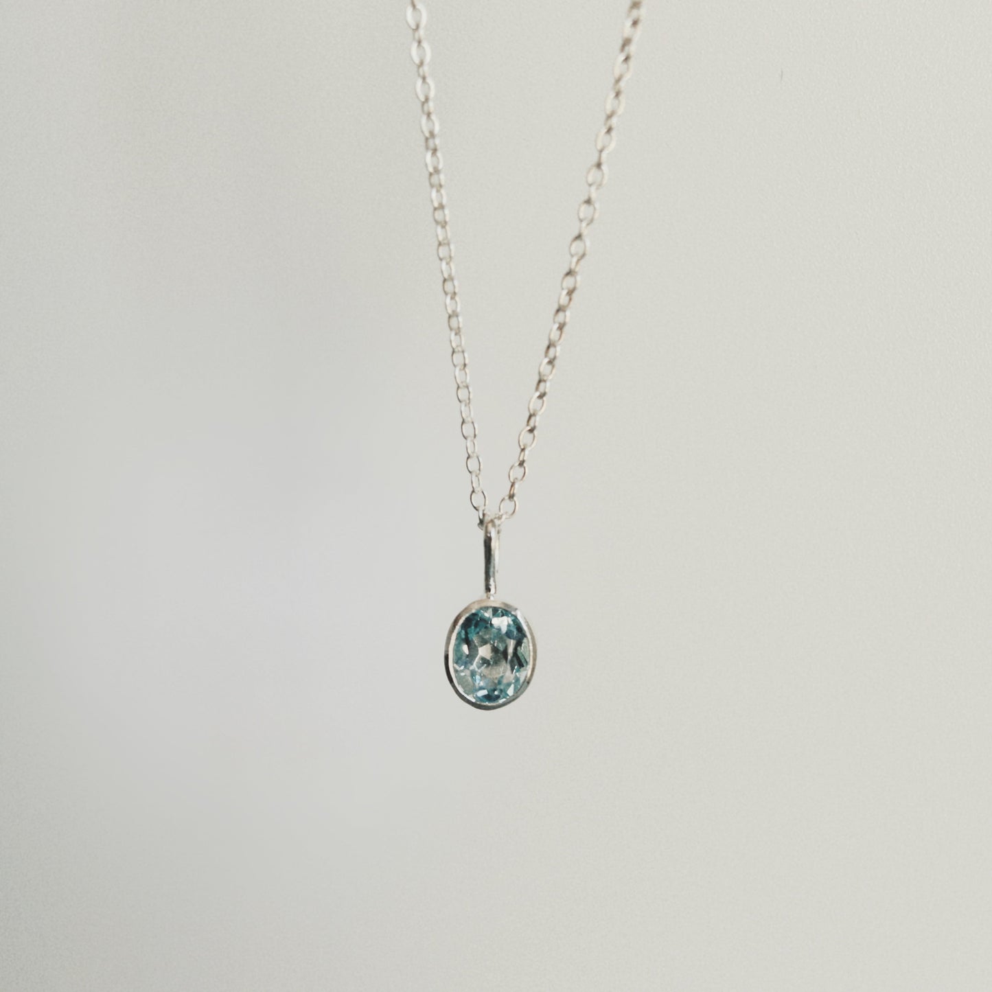 A delicate silver chain necklace with a small oval pendant featuring a light blue and white pattern, suspended against a plain light gray background.