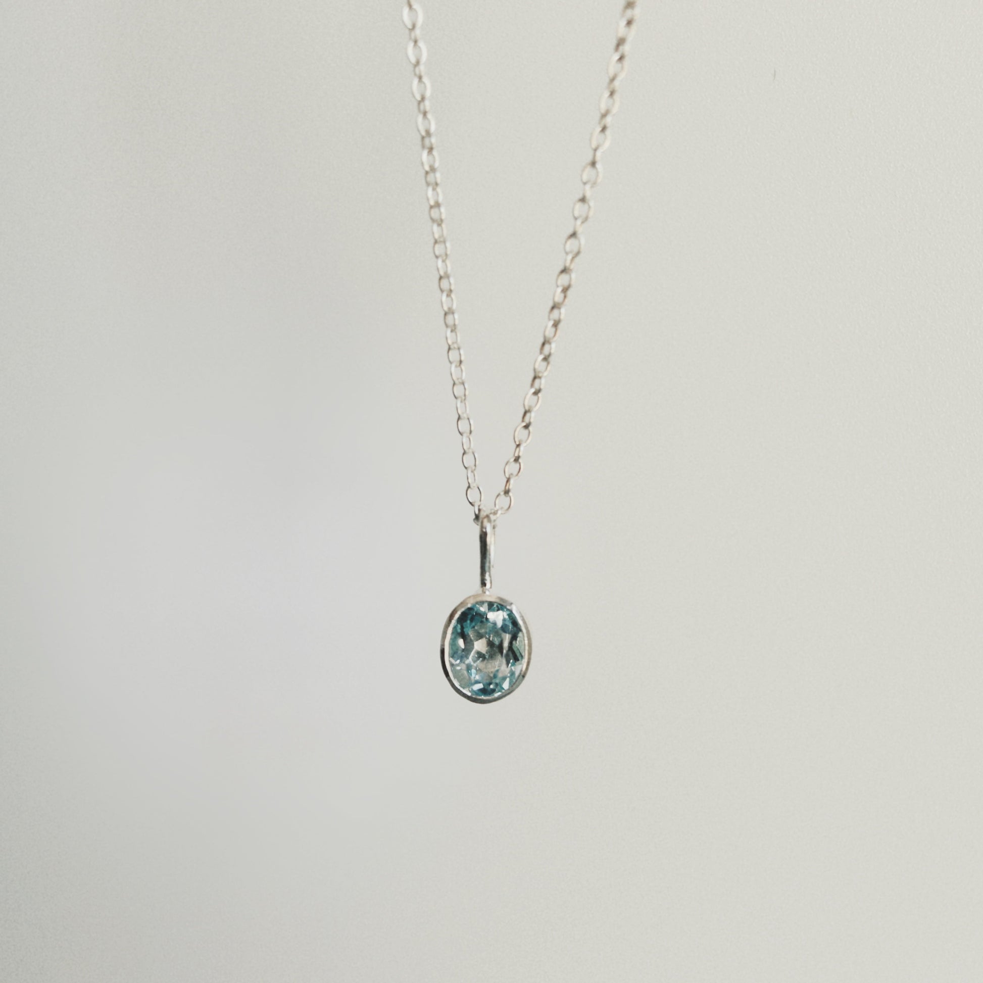 A delicate silver chain necklace with a small oval pendant featuring a light blue and white pattern, suspended against a plain light gray background.
