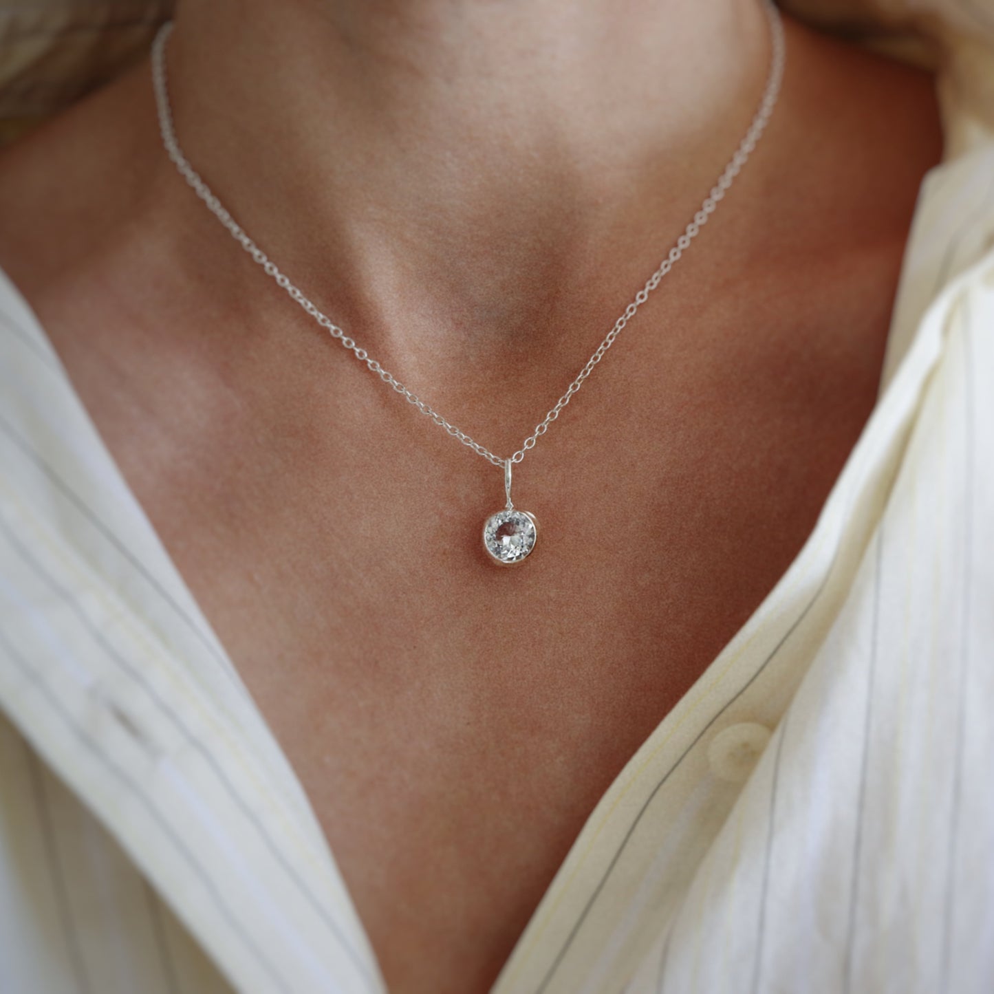 A close-up of a delicate silver chain necklace with a small round bezel-set clear gemstone pendant, worn by a model in a cream-colored shirt.