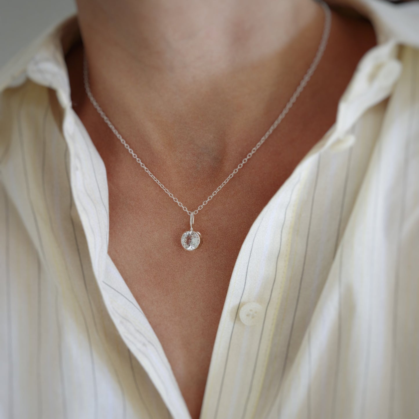 A close-up of a person wearing a delicate silver chain necklace with a small round clear pendant, styled with an open cream-colored button-down shirt.