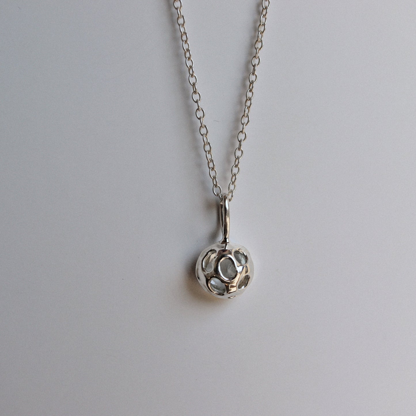 A silver pendant necklace featuring a small, textured circular charm with a central cutout, suspended from a delicate silver chain against a plain light gray background.