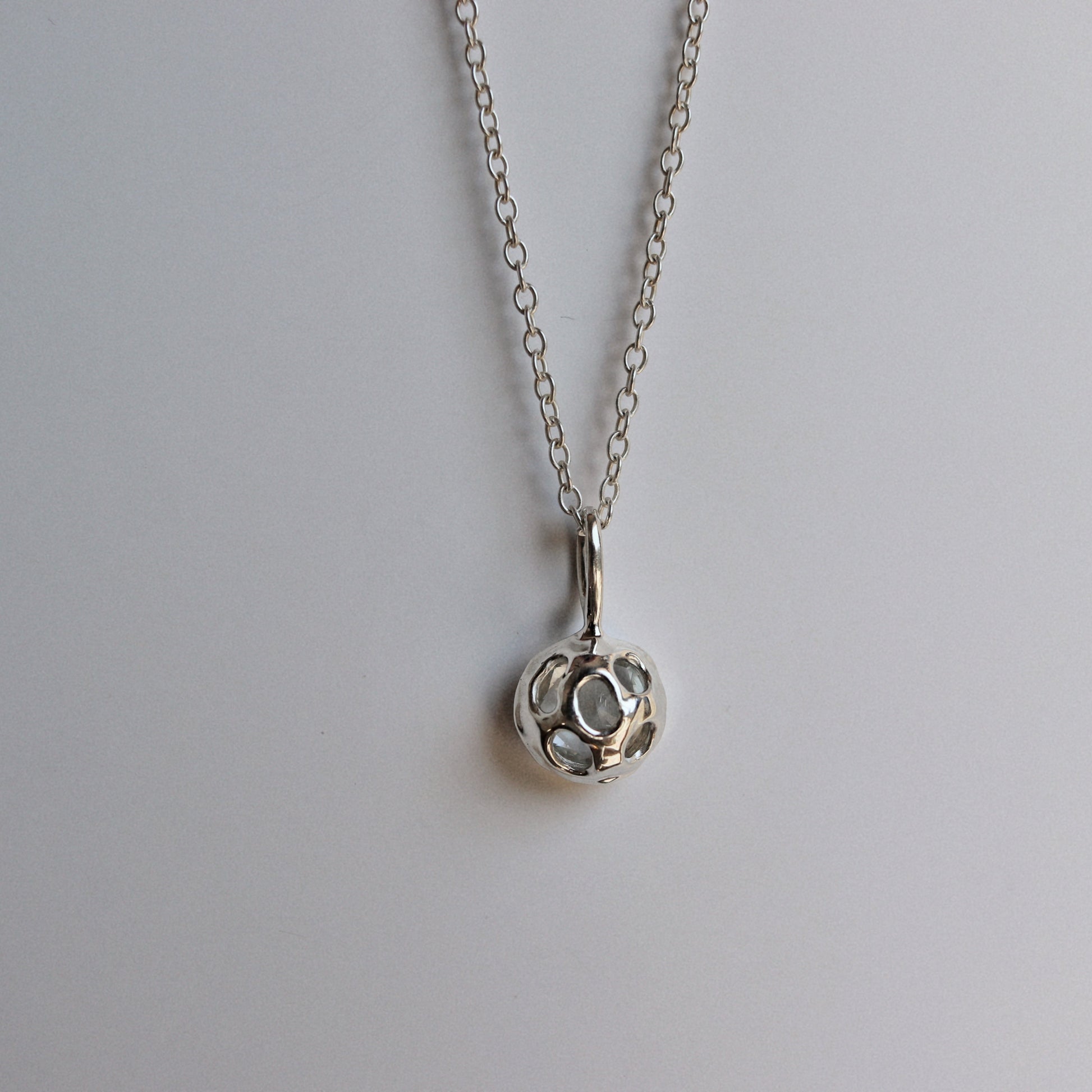 A silver pendant necklace featuring a small, textured circular charm with a central cutout, suspended from a delicate silver chain against a plain light gray background.
