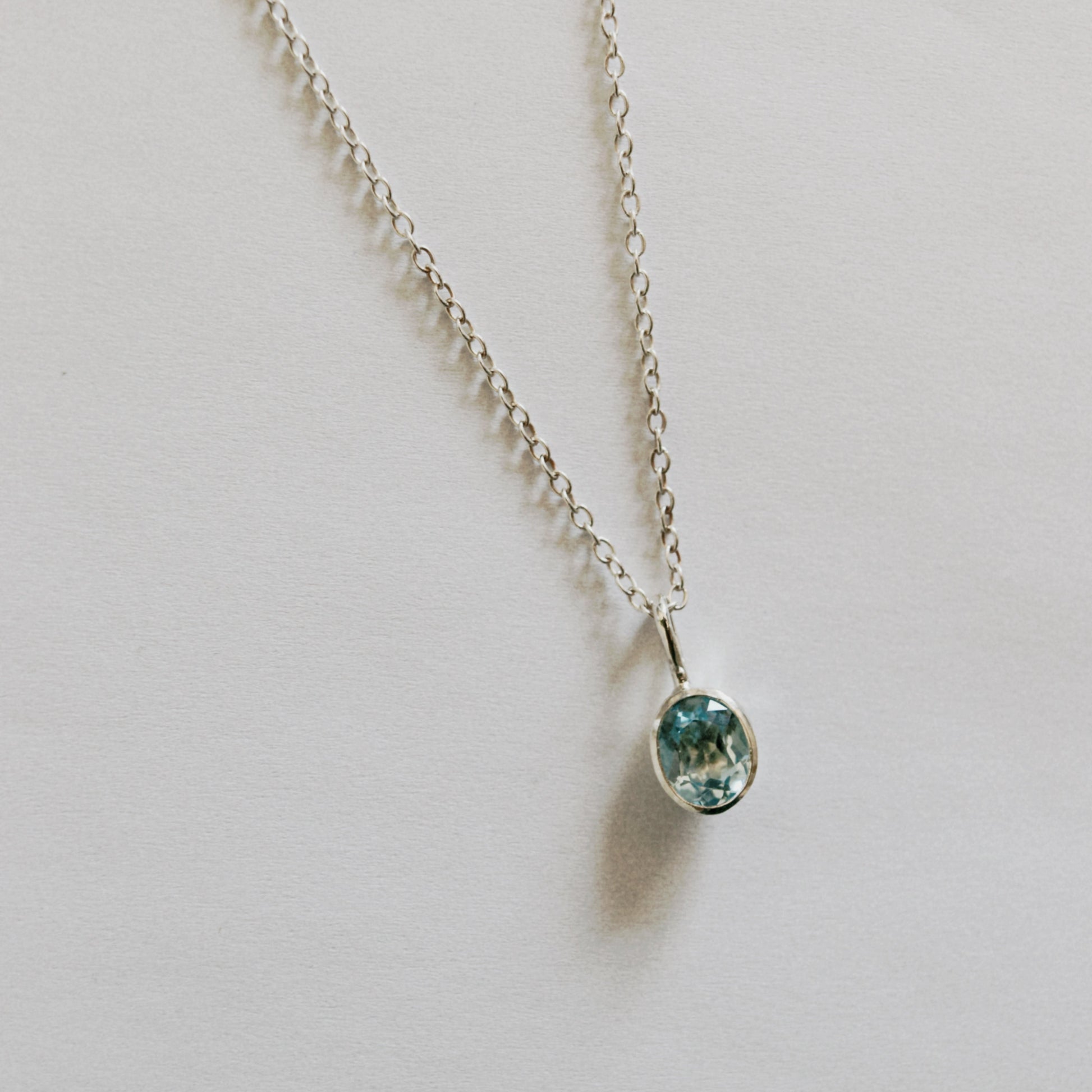 A delicate silver chain necklace with a small oval bezel pendant containing a light blue-green gemstone, displayed on a plain light gray background.