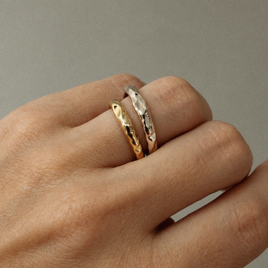 Two slim textured metal rings, one gold-toned and one silver-toned, worn on a person's fingers against a neutral gray background.