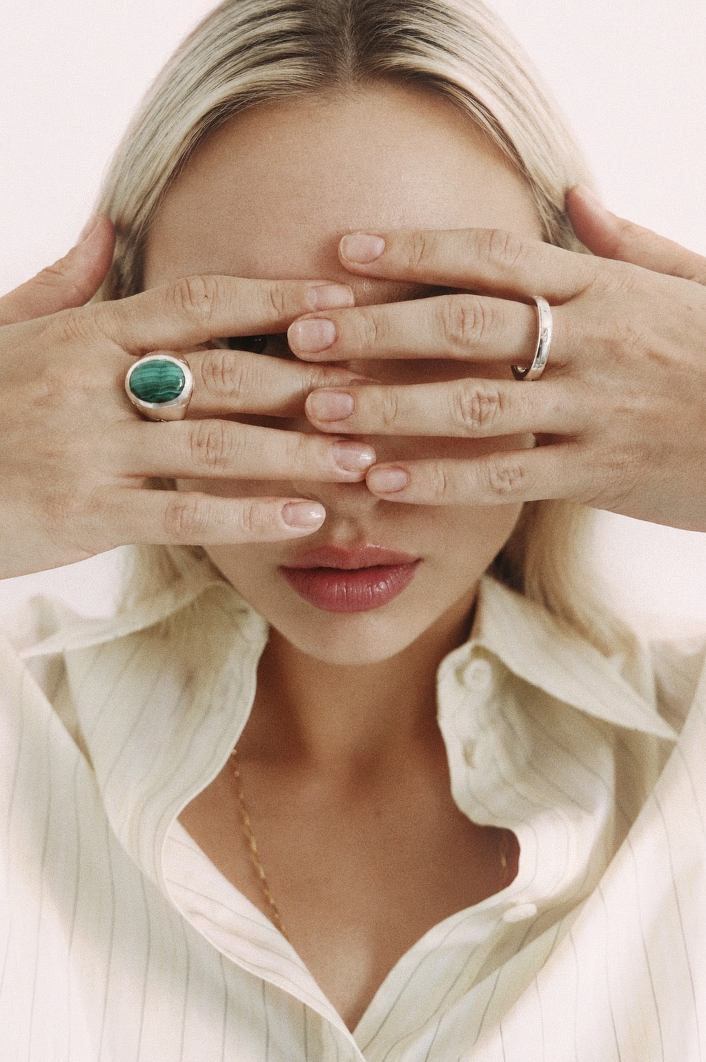 A close-up lifestyle shot of a woman with blonde hair covering her eyes with her hands, showcasing two rings: a prominent silver ring with a green oval stone on her left hand, and a thin silver band on her right hand.
