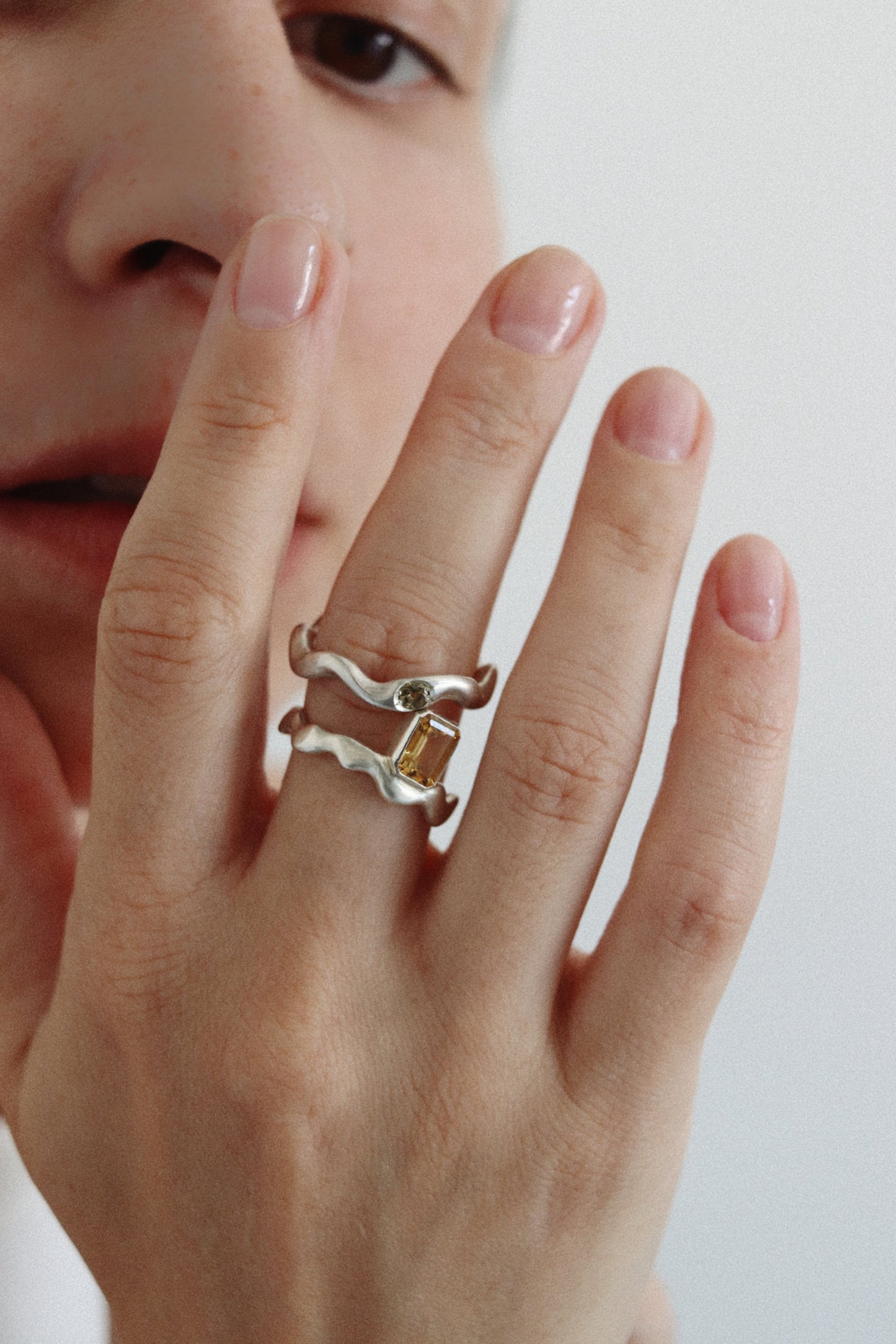 A close-up of a person's hand wearing a double-band ring with wavy silver and gold-toned metal bands, featuring a small green stone and a central yellow crystal.