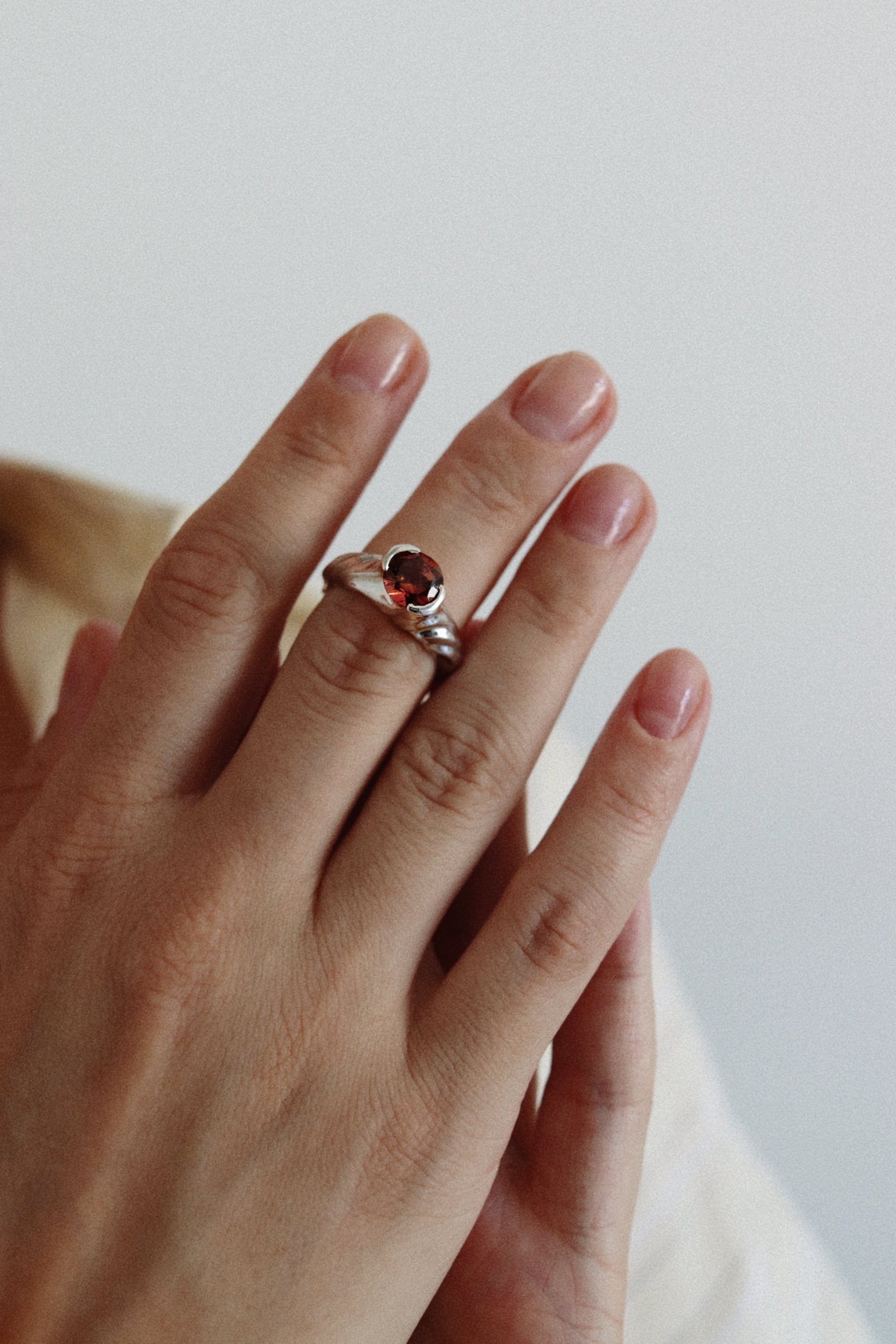A close-up of a hand wearing a silver ring with a round red gemstone, set against a soft, light gray background.