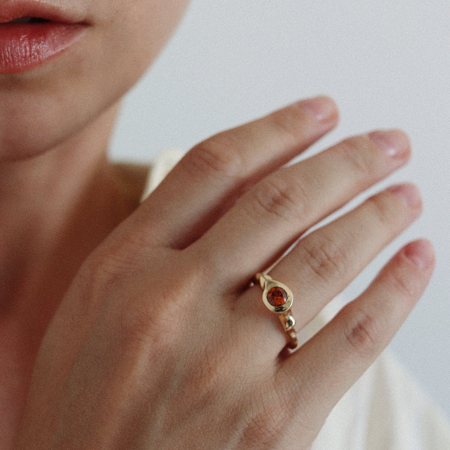A close-up lifestyle shot of a woman's hand wearing a delicate gold ring with a central round amber gemstone, against a soft, light gray background.