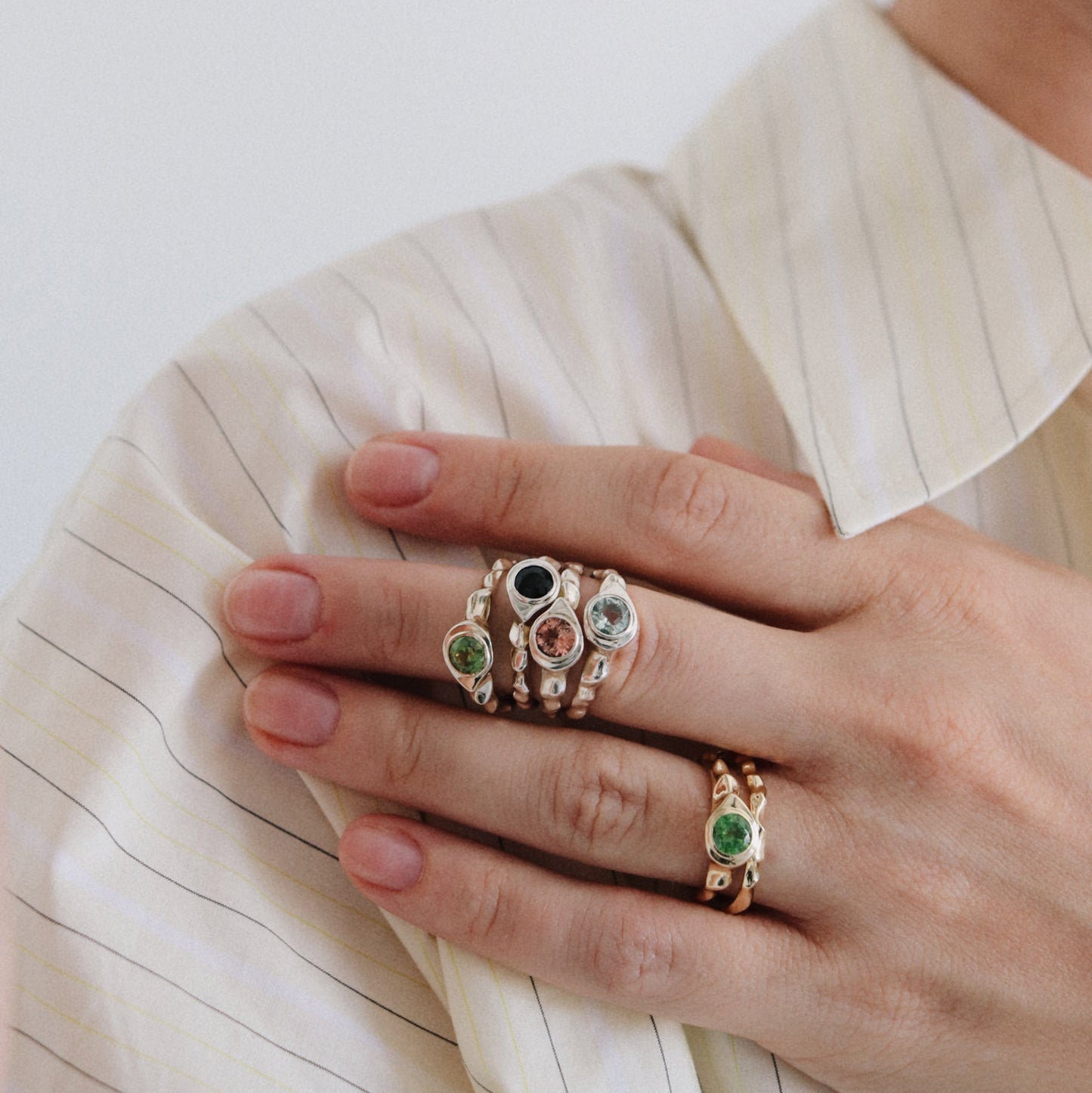 A close-up of a person's hand wearing multiple rings, including a gold ring with a green emerald and a stack of rings with colorful gemstones, set against a cream pinstriped shirt.