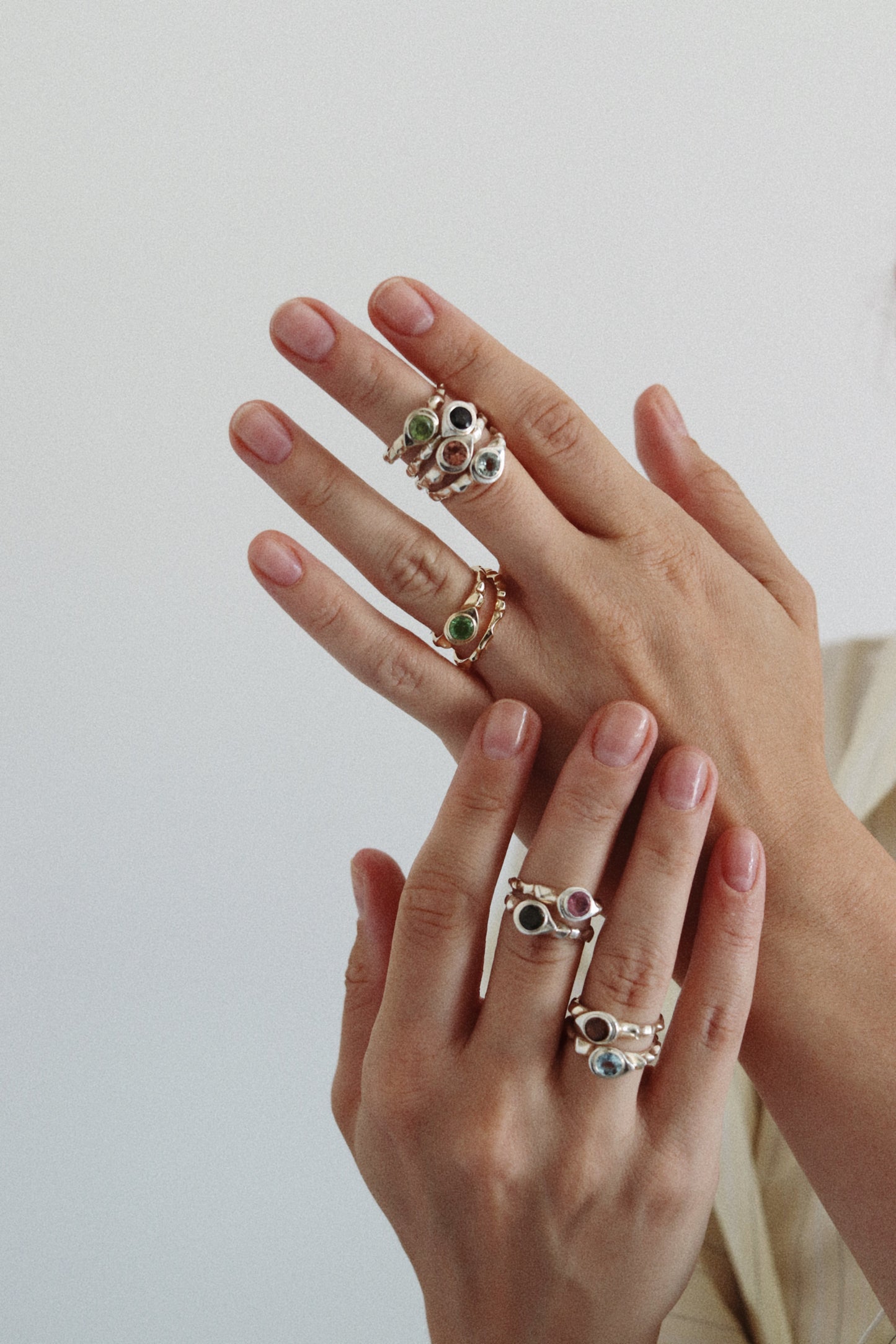 A close-up of two hands wearing multiple delicate rings with small green and blue stones set in silver-toned metal, against a soft light gray background.