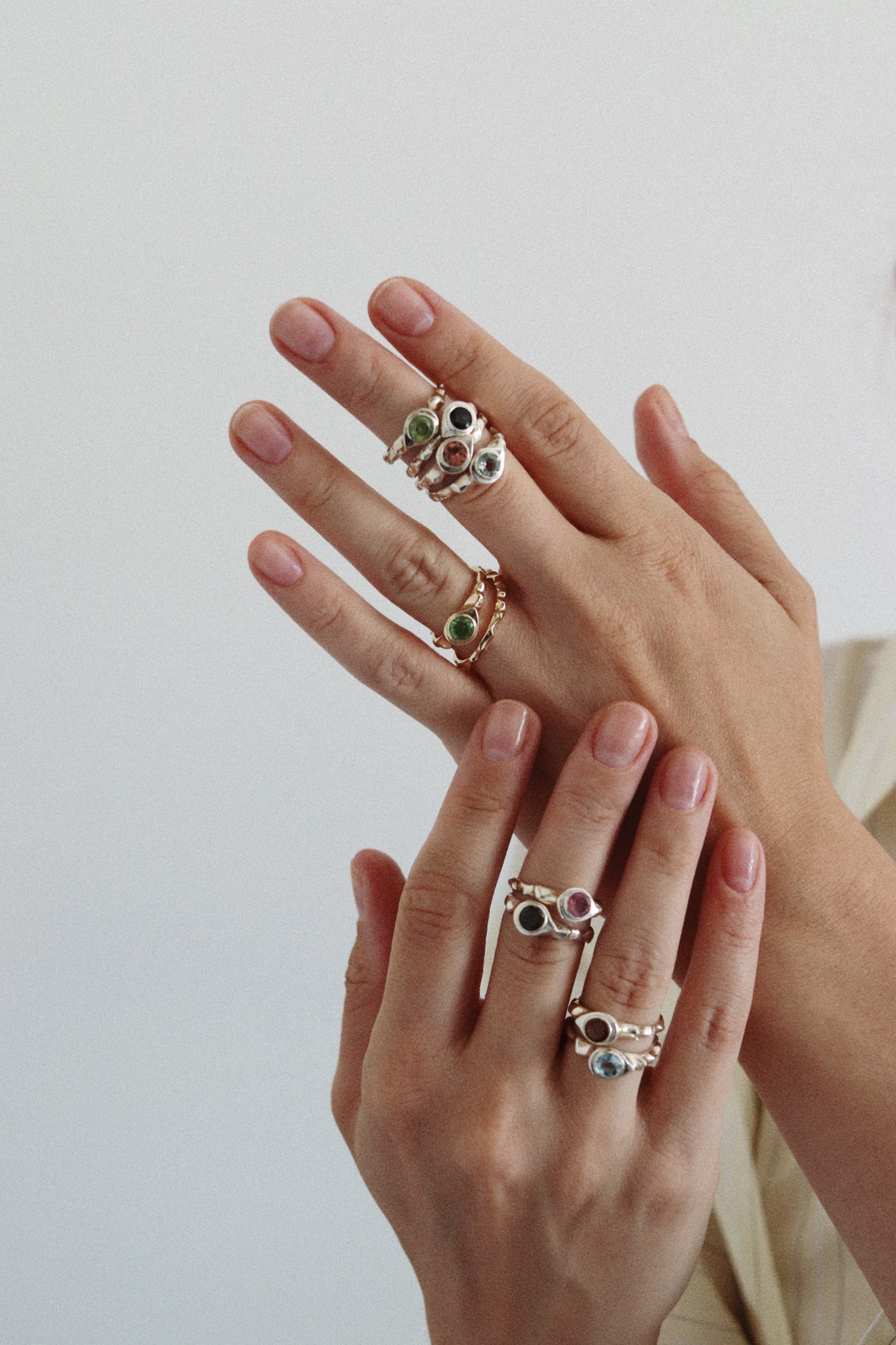 A close-up of two hands wearing multiple delicate rings with small green and blue stones set in silver-toned metal, against a soft light gray background.