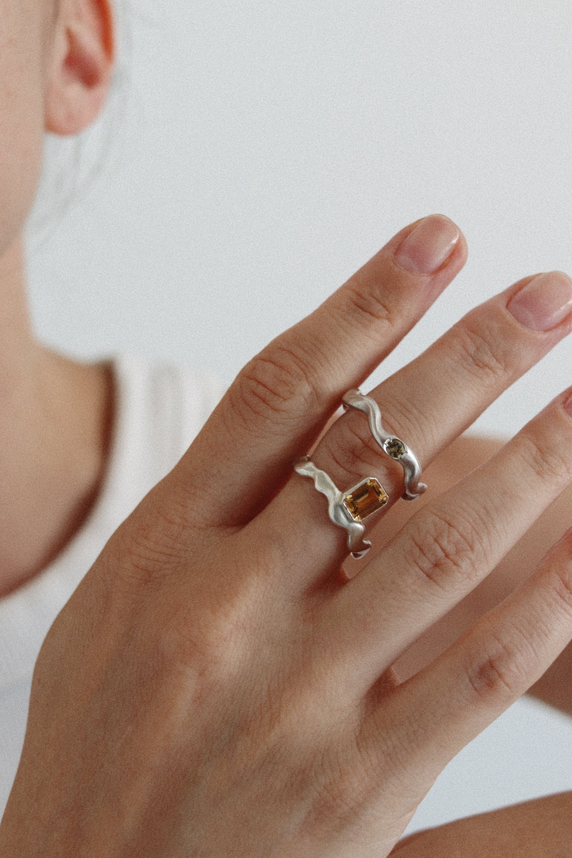 A close-up of a person's hand wearing a silver wavy band ring with a small oval amber gemstone, against a soft light gray background.