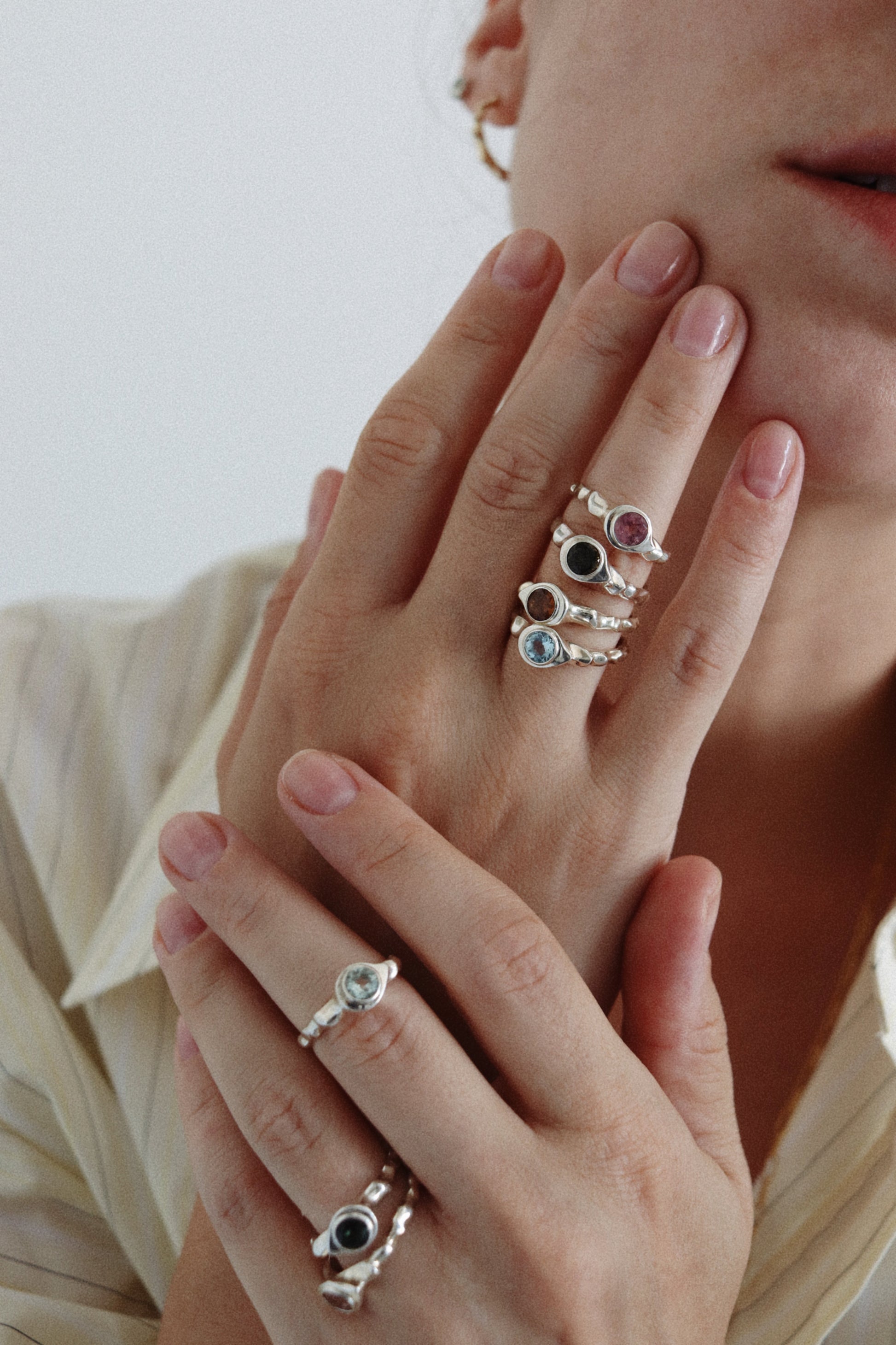 A close-up of a person's hands wearing multiple silver rings with colorful gemstones, including pink, blue, and dark green stones, against a light neutral background.