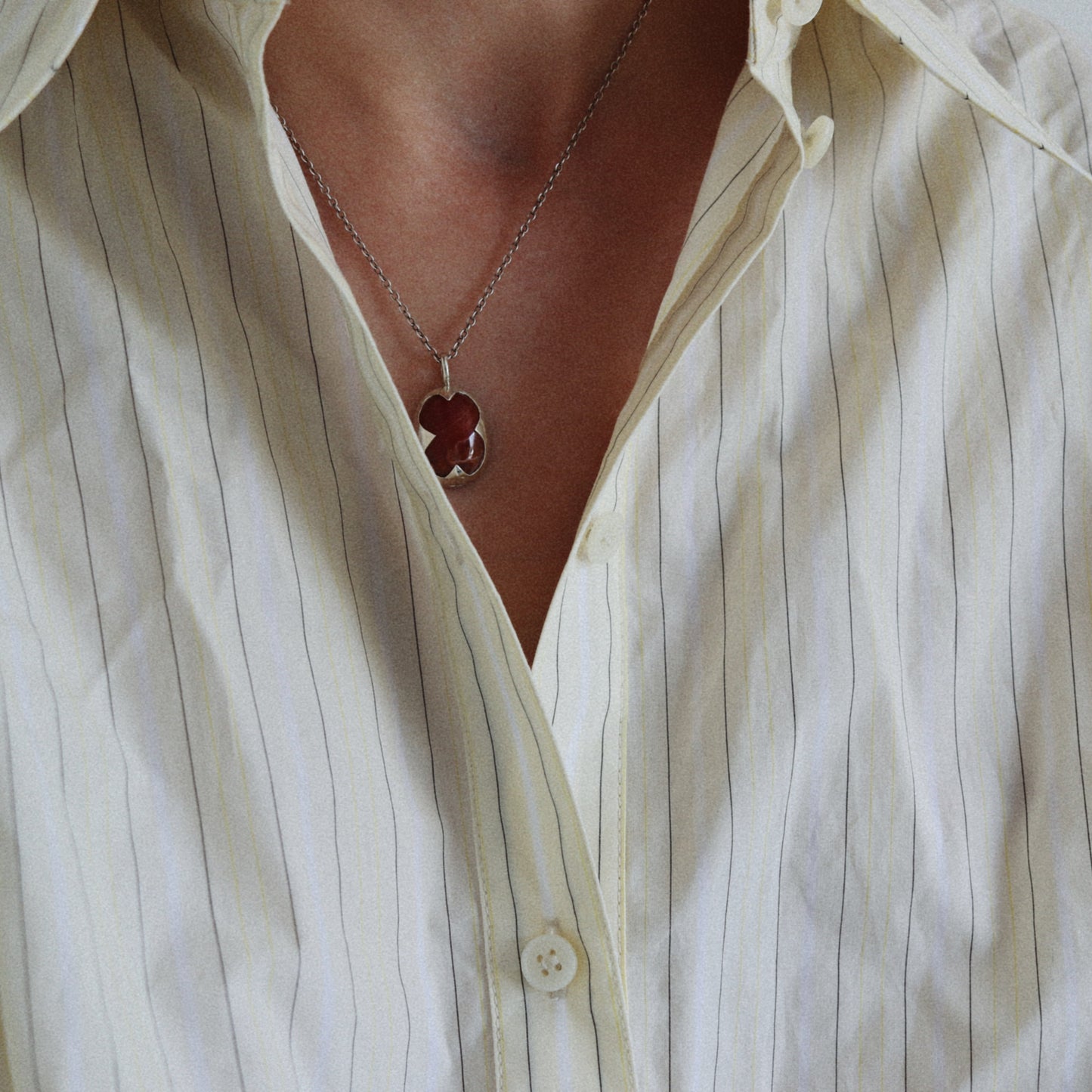 A close-up of a person wearing a cream-colored pinstriped button-up shirt and a silver chain necklace with a dark red clover-shaped pendant.