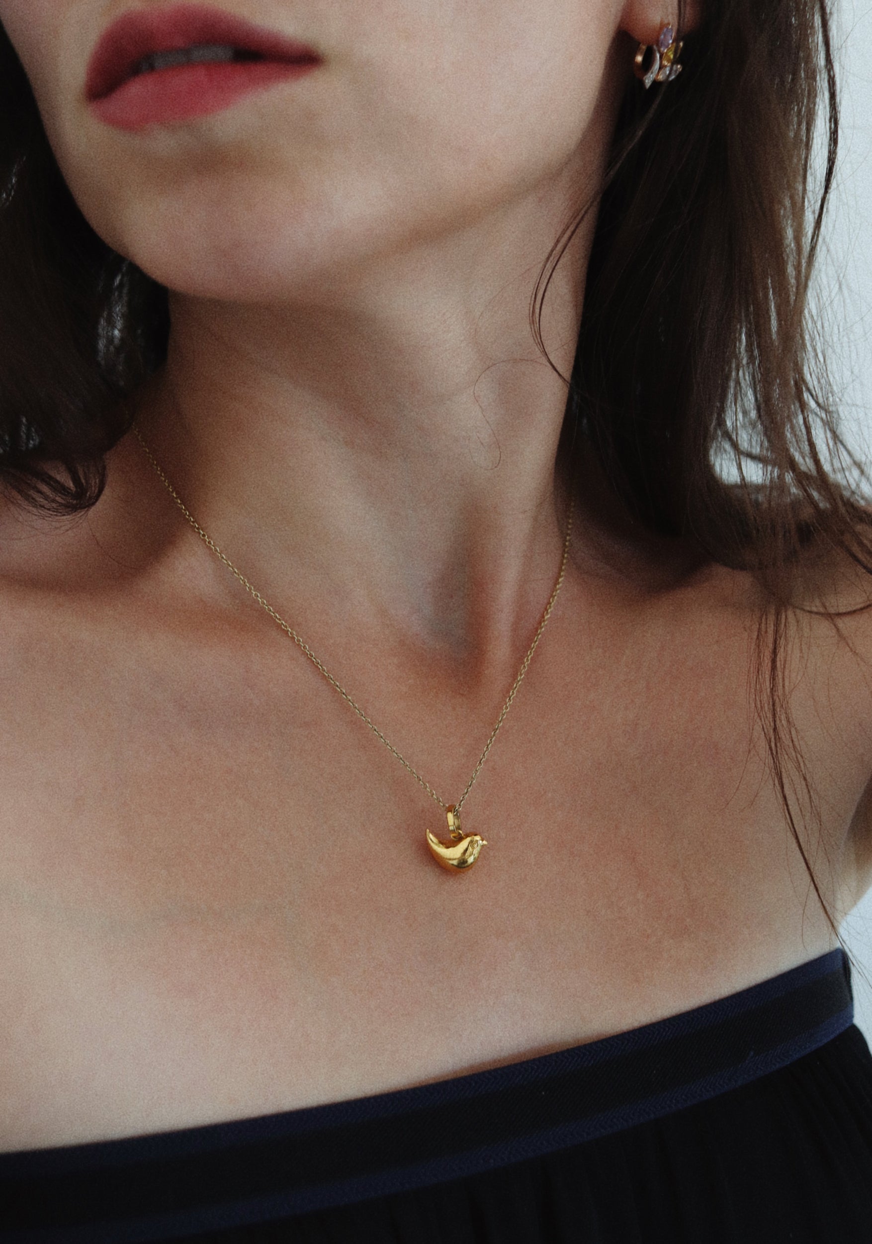 A close-up lifestyle shot of a woman wearing a delicate gold necklace with a small bird pendant, paired with a dark navy off-the-shoulder top.