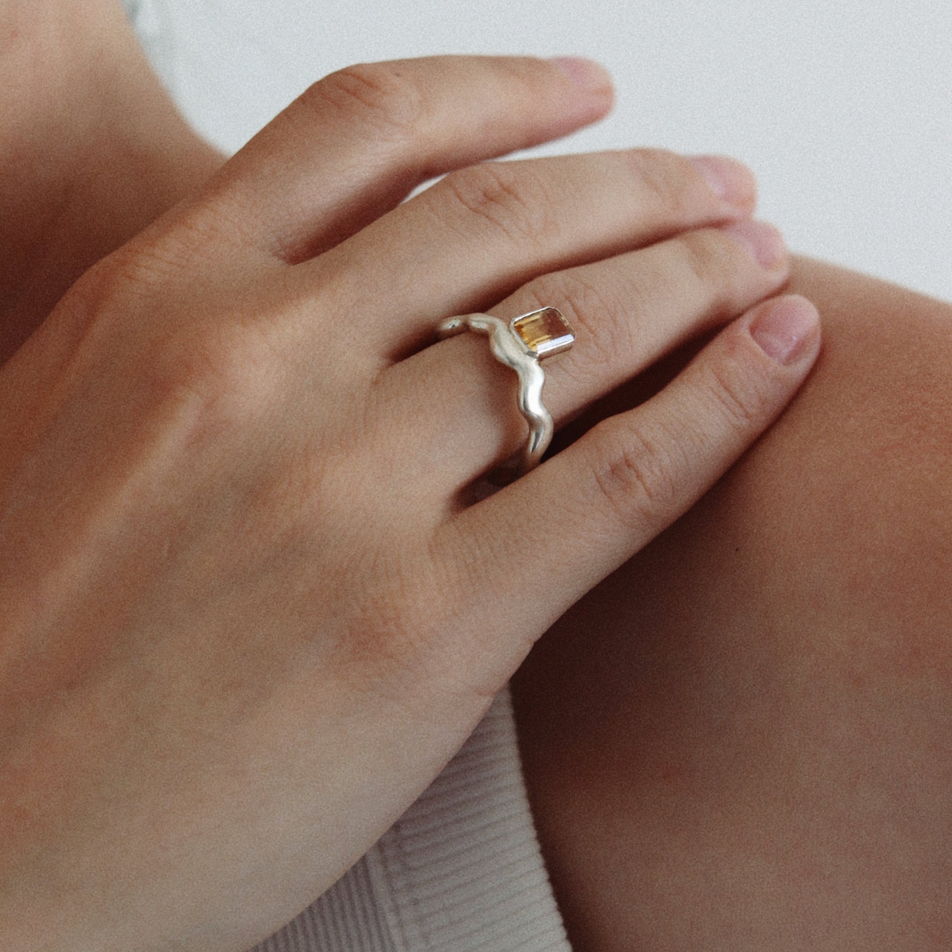 A close-up of a person's hand wearing a delicate silver ring with a small gold heart-shaped accent, resting on their shoulder against a light gray background.