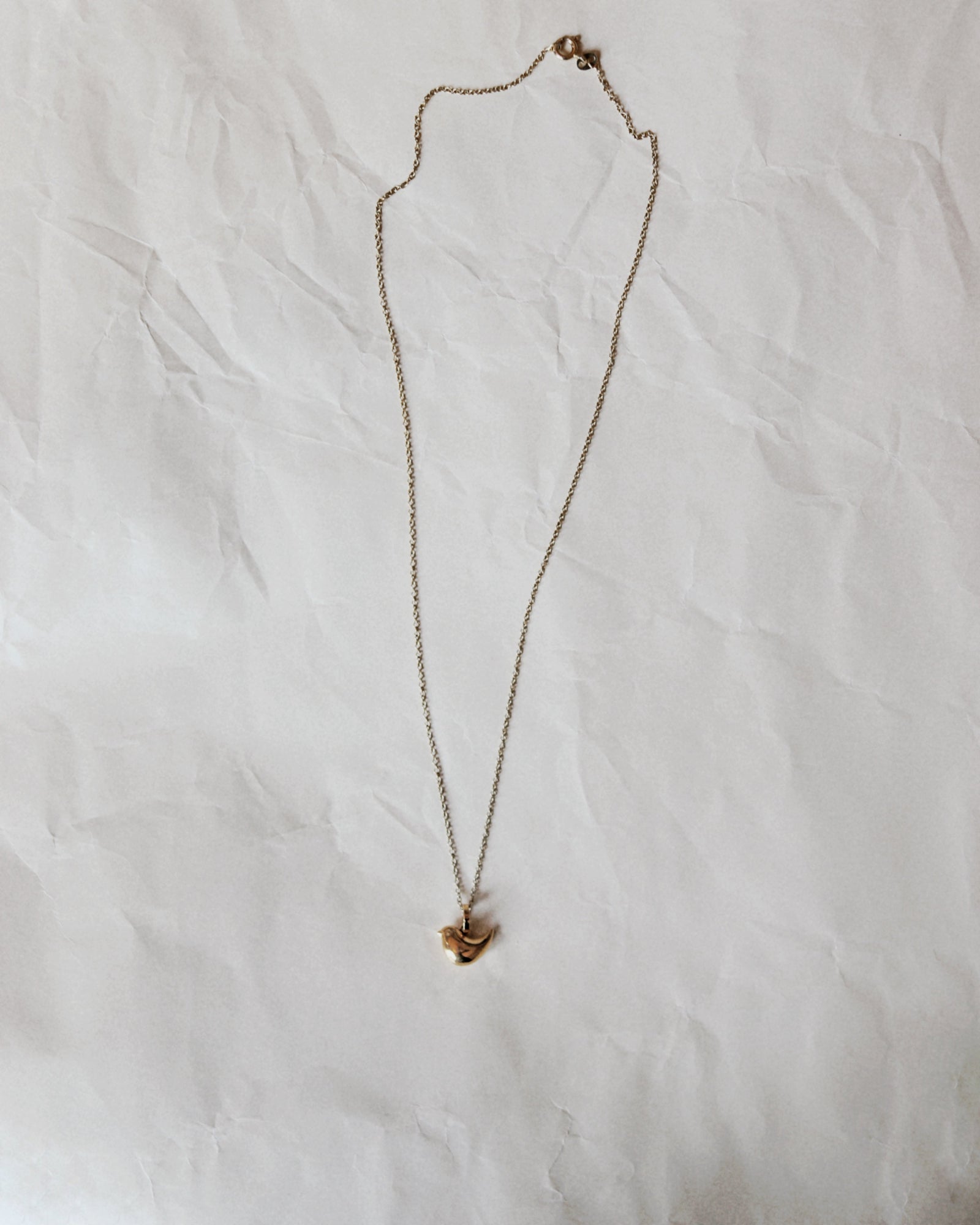 A delicate gold chain necklace with a small gold pendant, laid flat on a wrinkled white paper background.