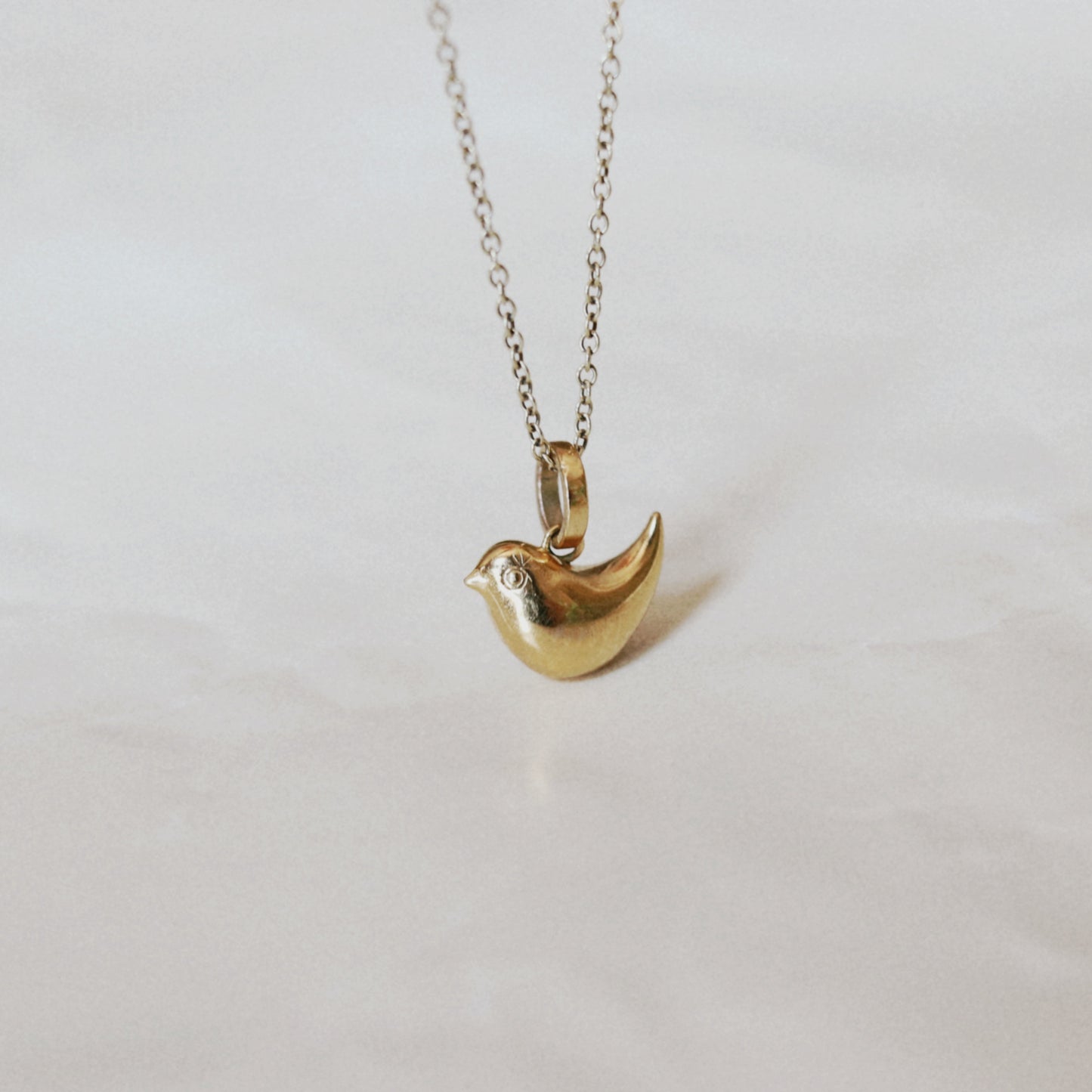 A gold bird-shaped pendant necklace on a delicate silver-toned chain, displayed against a soft, light beige fabric background.