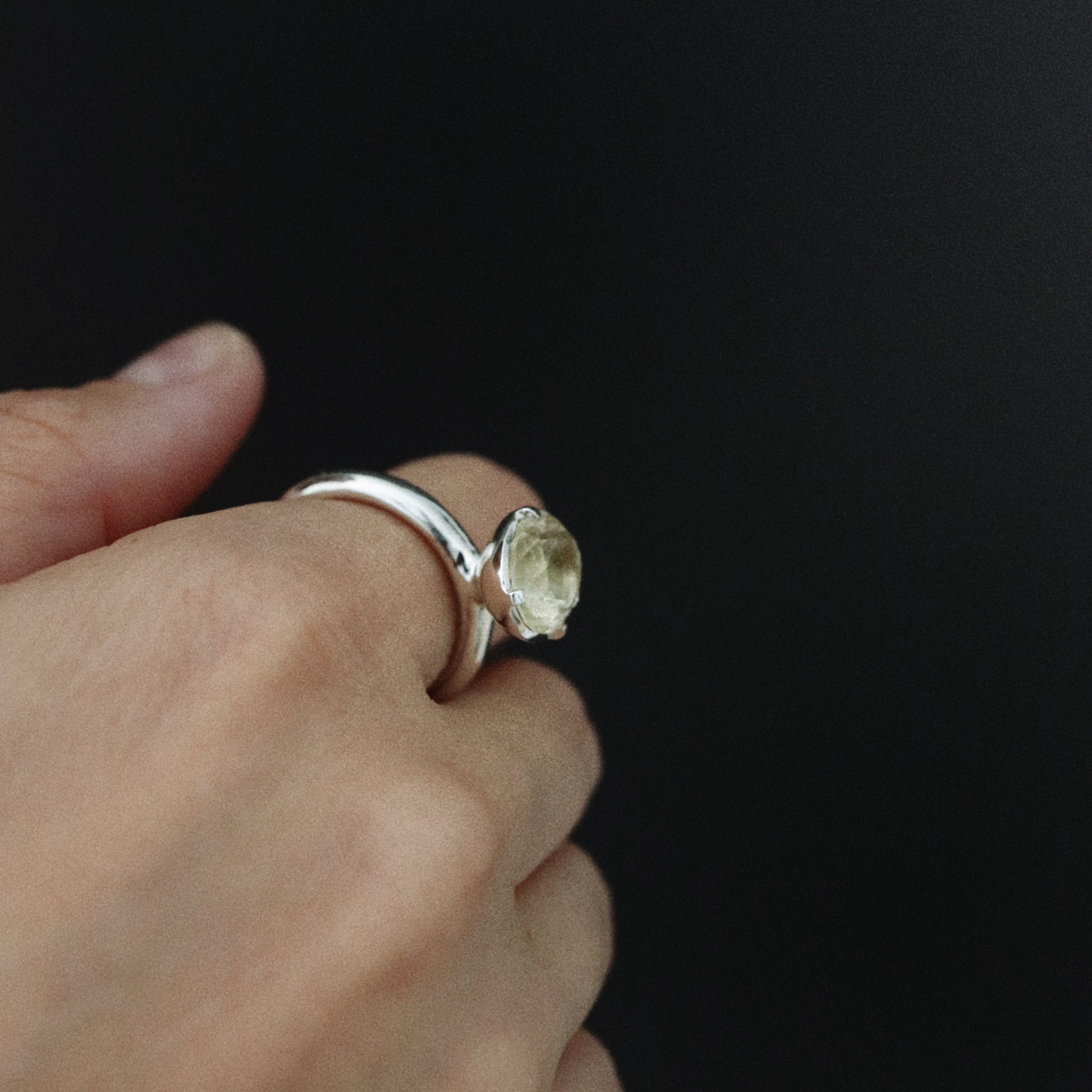 A silver ring with a round, bezel-set pale yellow gemstone worn on a person's finger, set against a solid black background.