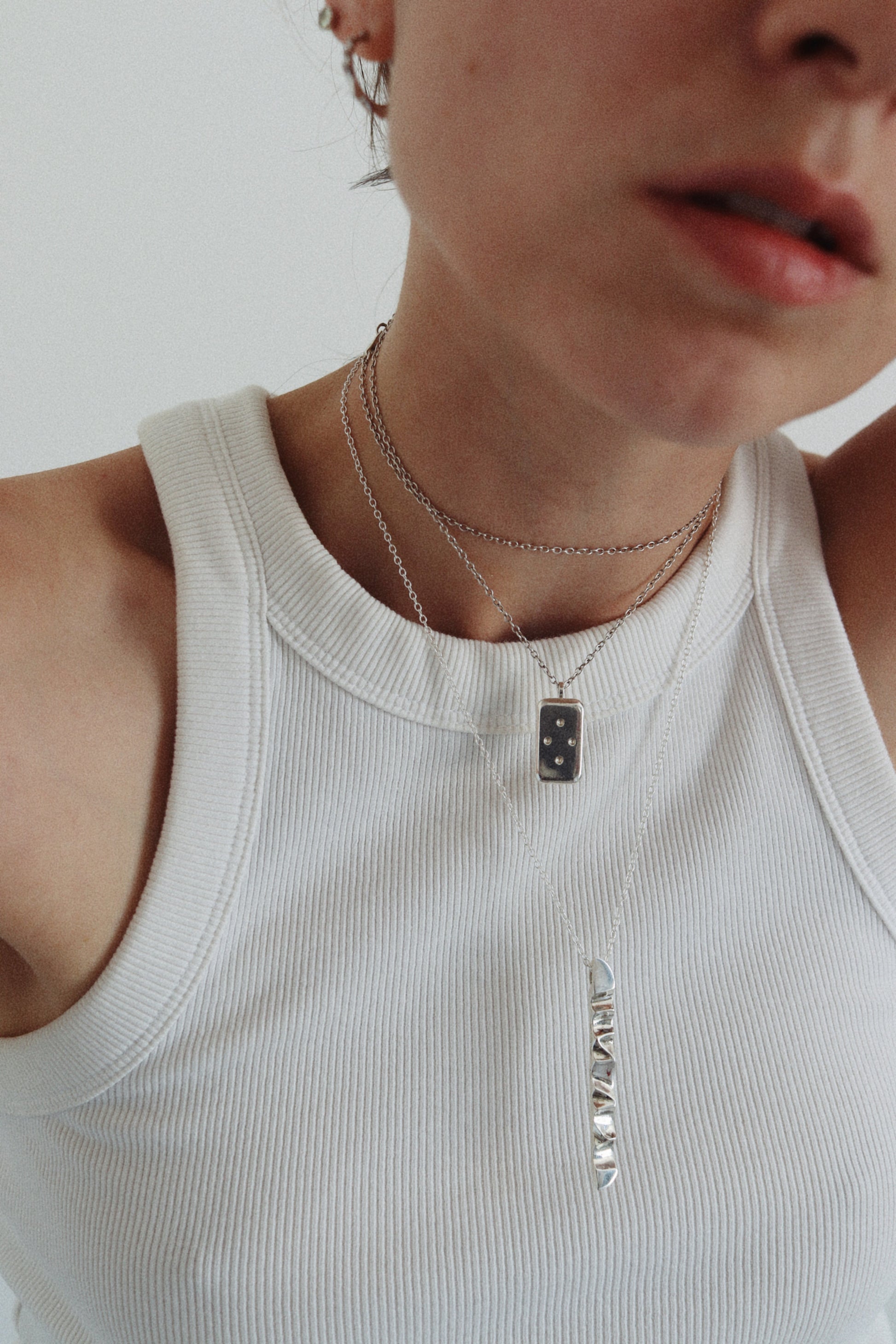 Close-up of a person wearing layered silver necklaces, including a small rectangular pendant and a vertical bar necklace, over a white ribbed tank top.