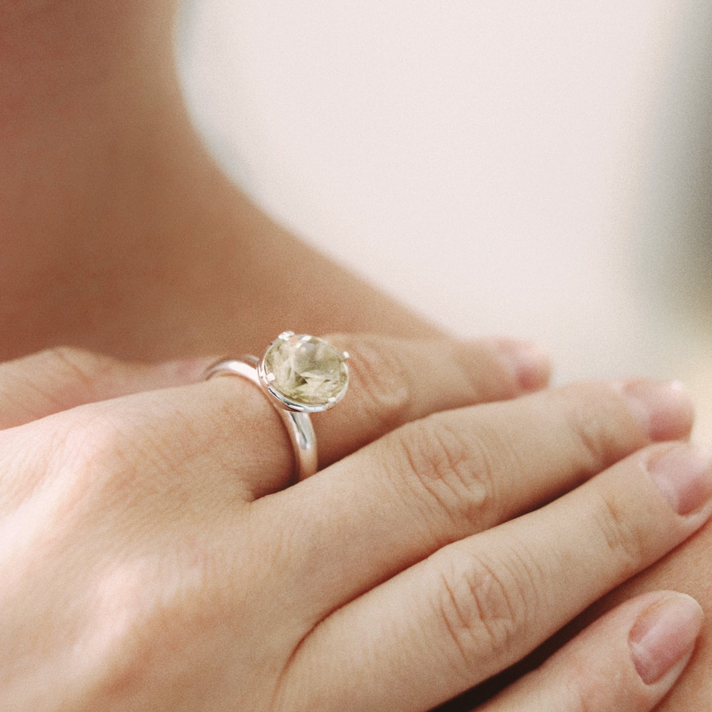 A close-up lifestyle shot of a silver ring with a round clear gemstone worn on a person's finger, with their hand resting on their lap against a soft, warm neutral background.