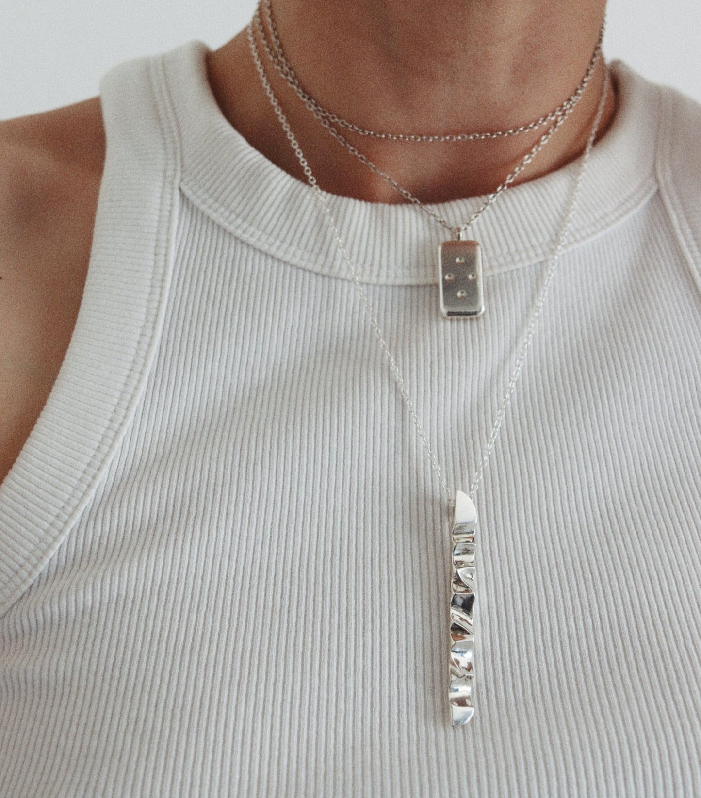 A close-up of a person wearing layered silver necklaces, including a long vertical bar pendant and a smaller rectangular pendant, over a ribbed white tank top.