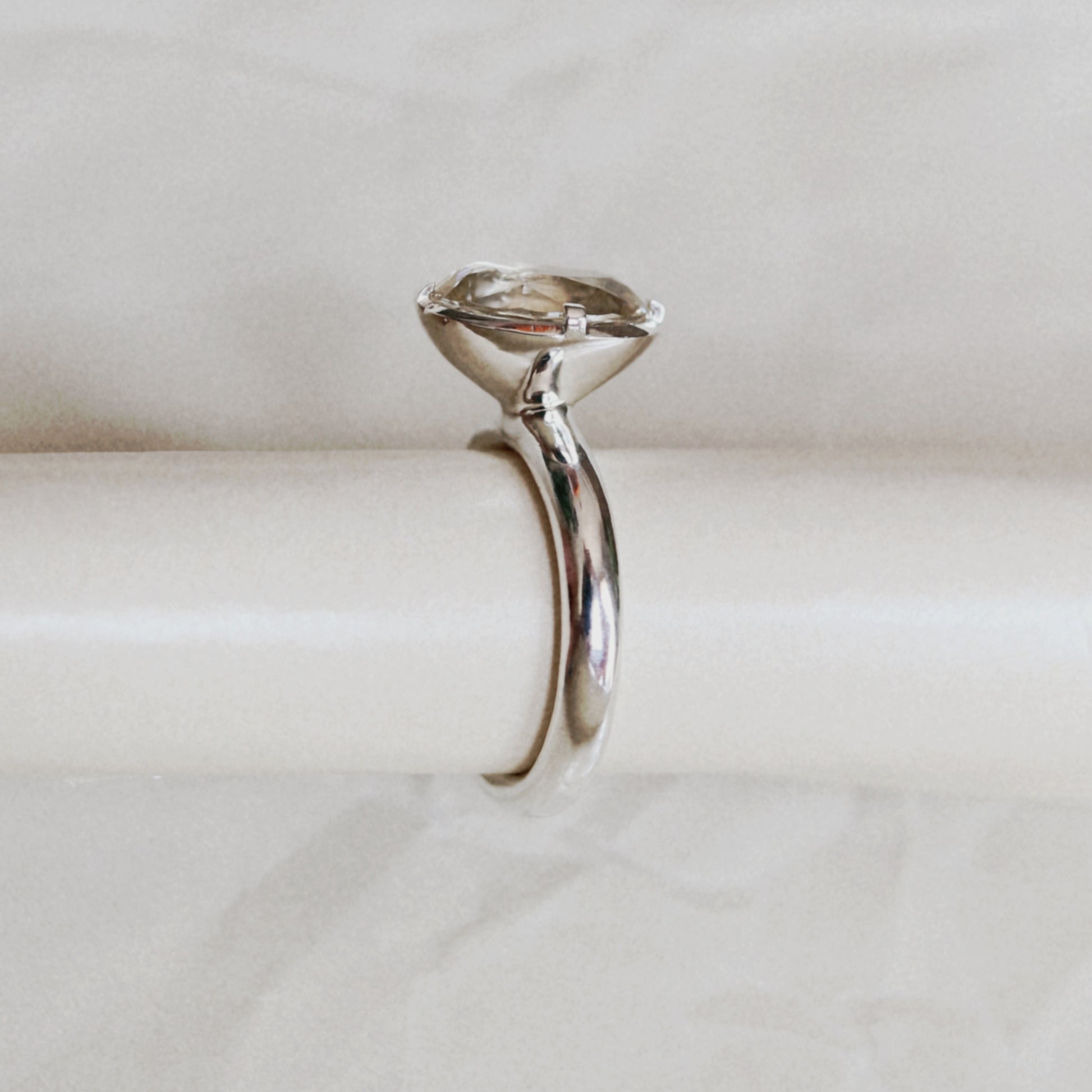 A polished silver-toned solitaire ring with a marquise-cut gemstone in a four-prong setting, displayed on a cream-colored cylindrical stand against a light marble background.