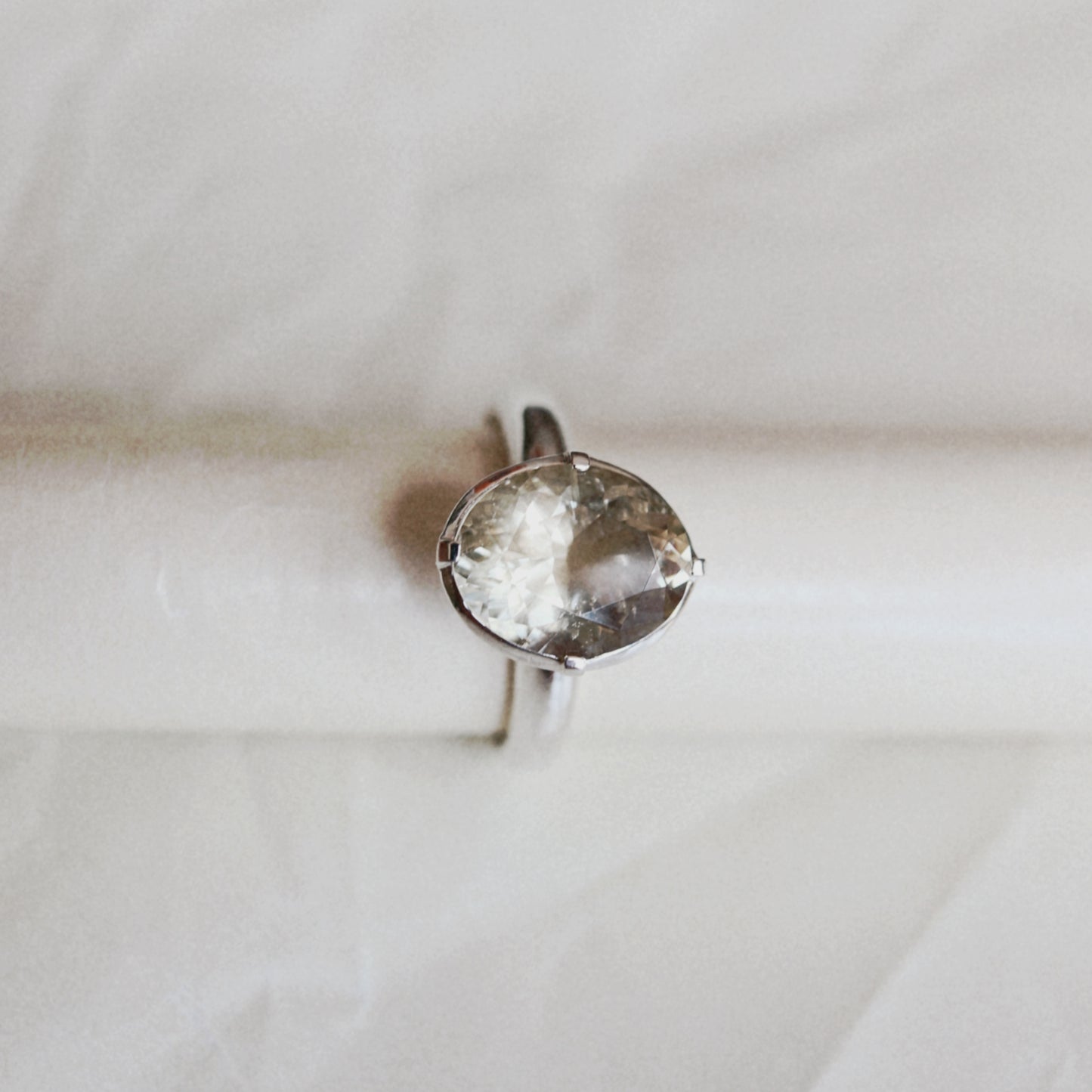 A silver ring with a large, oval, smoky clear gemstone, displayed on a white cylindrical ring stand against a soft, light gray fabric background.