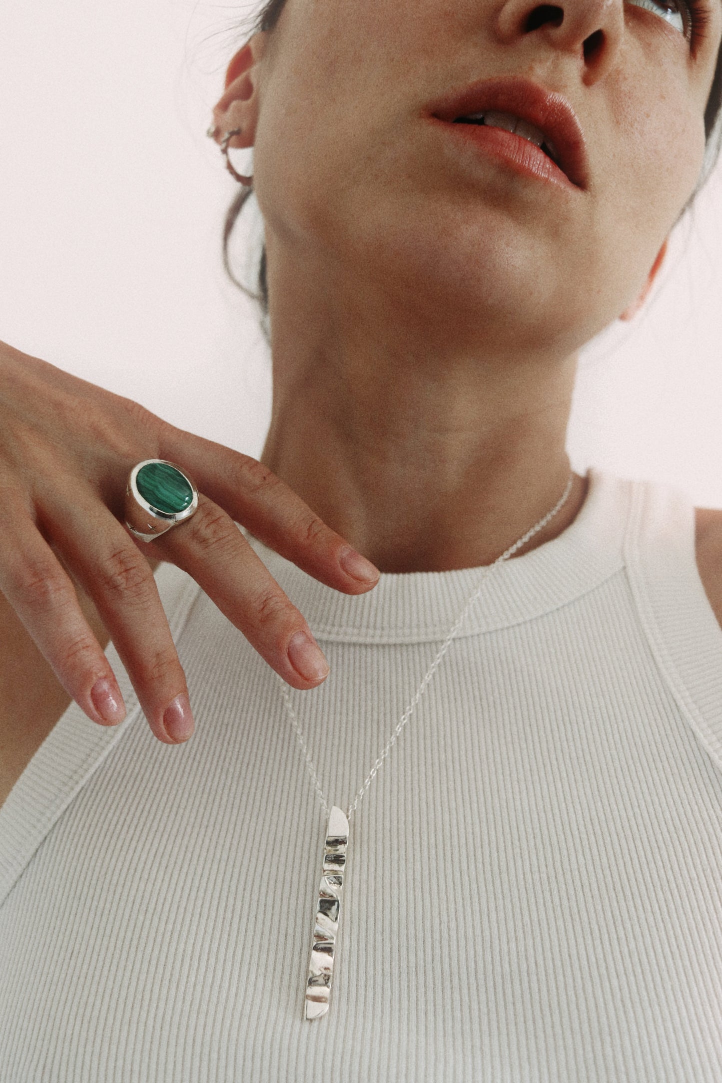 A close-up lifestyle shot of a model wearing a silver pendant necklace with clear stones and a silver ring with a green stone.