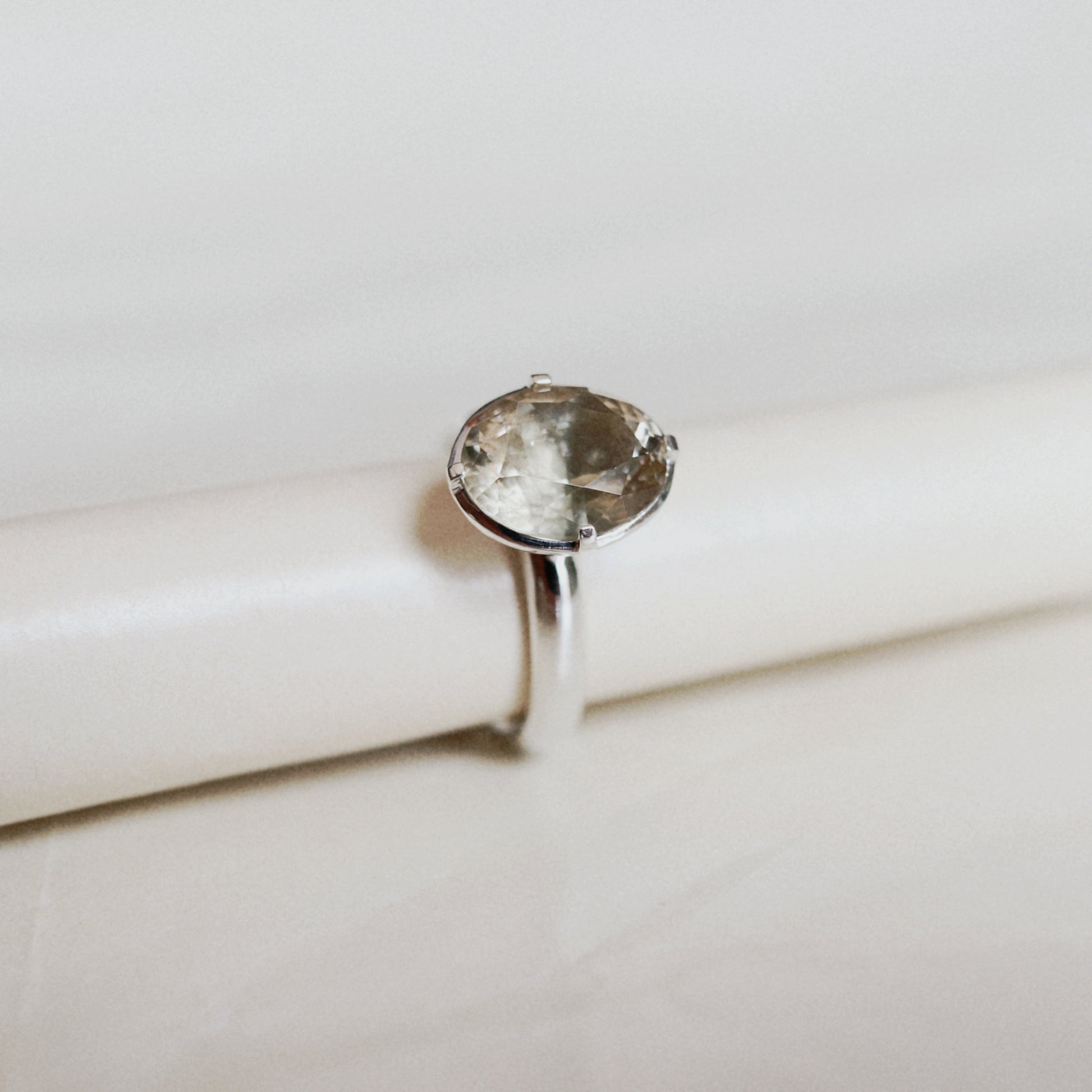 A silver ring with a large oval clear gemstone, displayed on a white cylindrical stand against a light beige background.