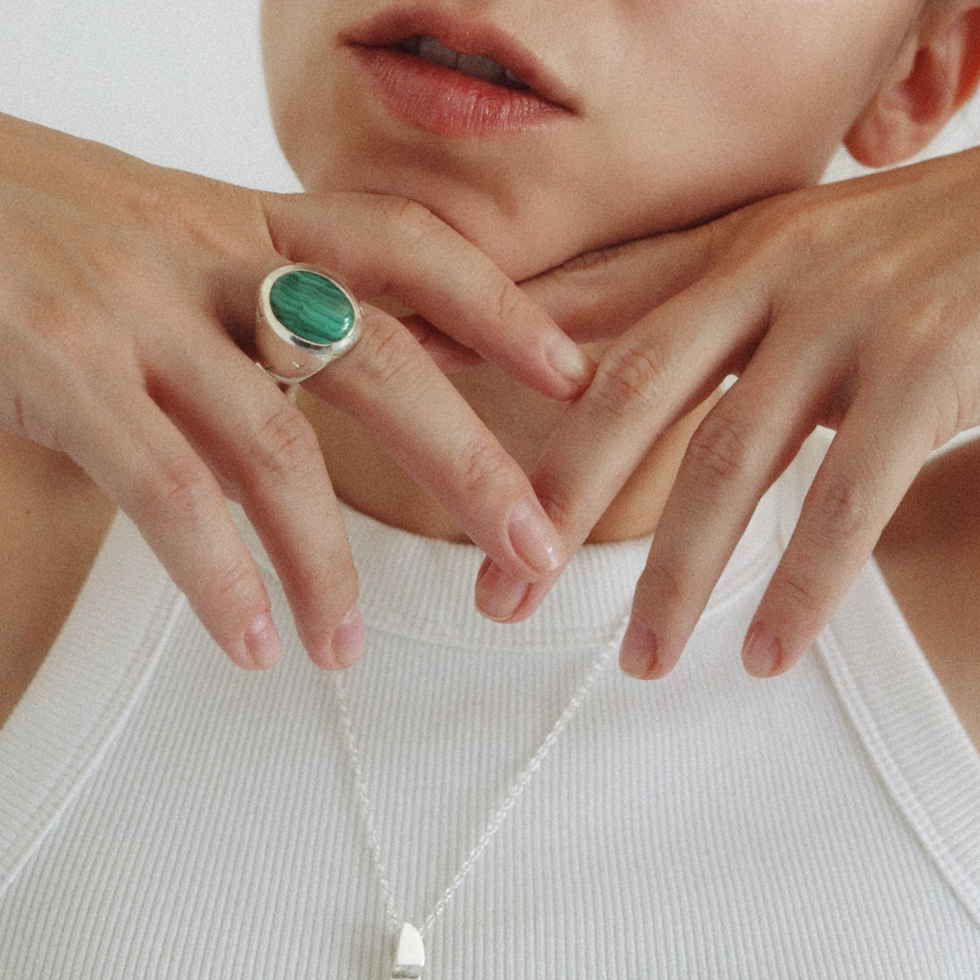 A close-up lifestyle shot of a woman wearing a silver ring with a round green stone, resting her chin on her hands against a plain white background.