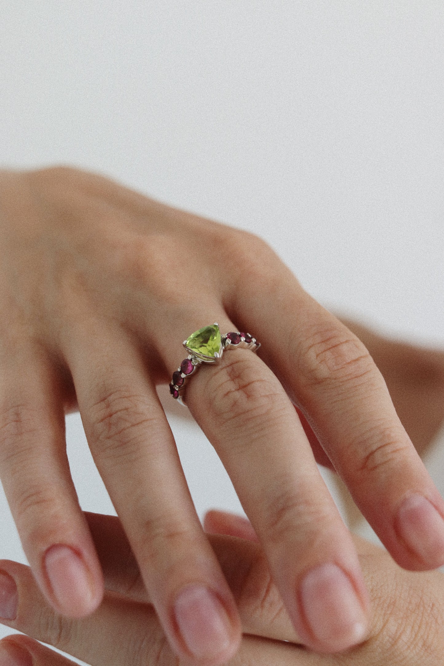 A close-up of a hand wearing a silver ring with a green heart-shaped gemstone and small pinkish-purple accent stones.