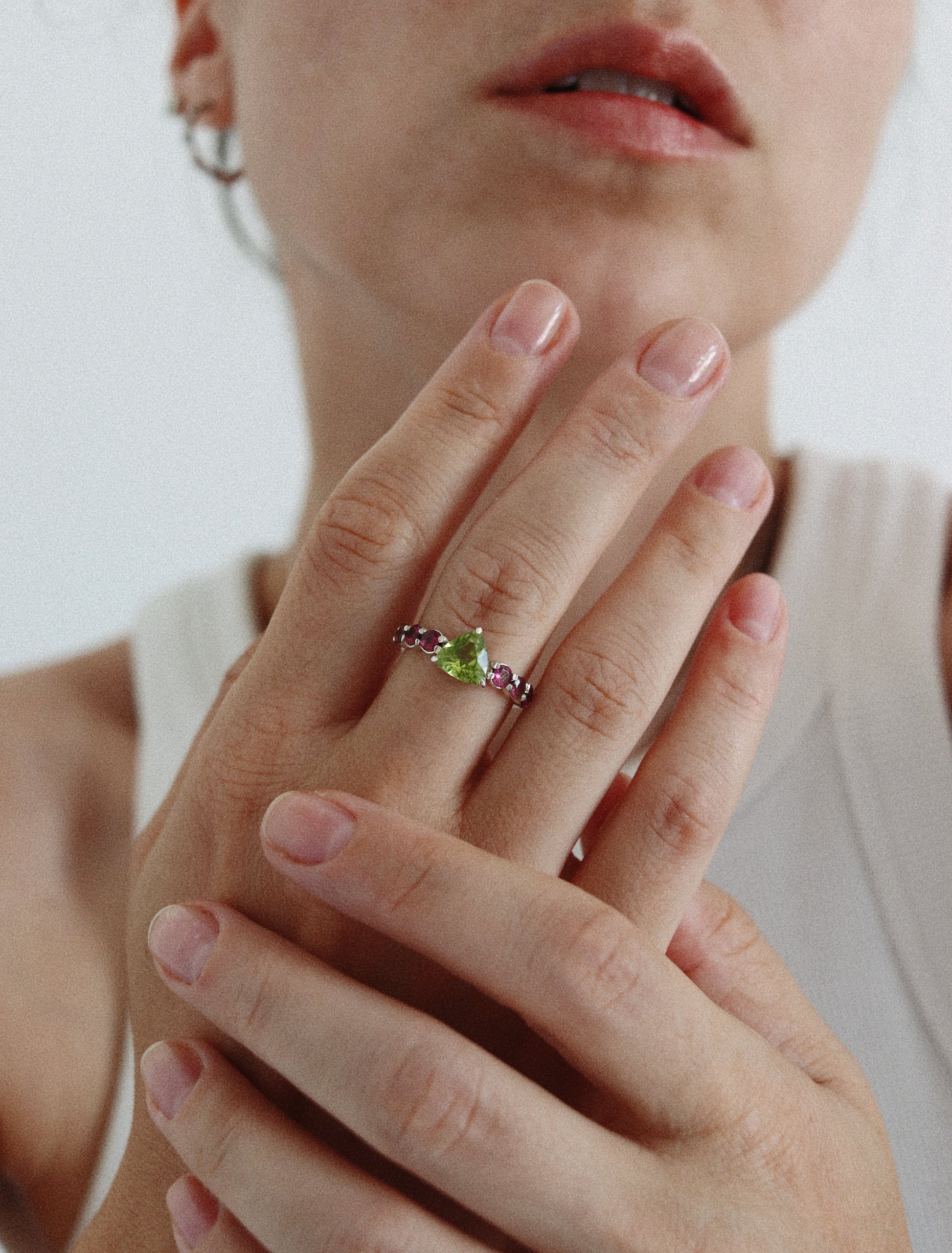 A close-up of a person's hands and lower face wearing a delicate silver ring with a small green marquise-cut gemstone and tiny pink accent stones.