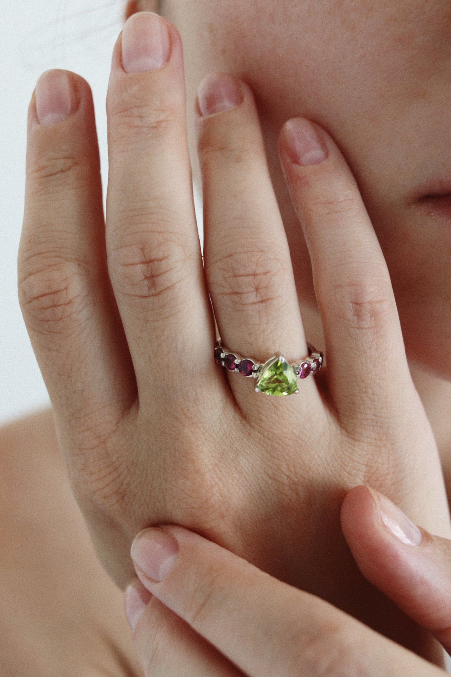 A close-up of a hand wearing a silver ring with a green triangular center stone and small purple accent stones on either side of the band.
