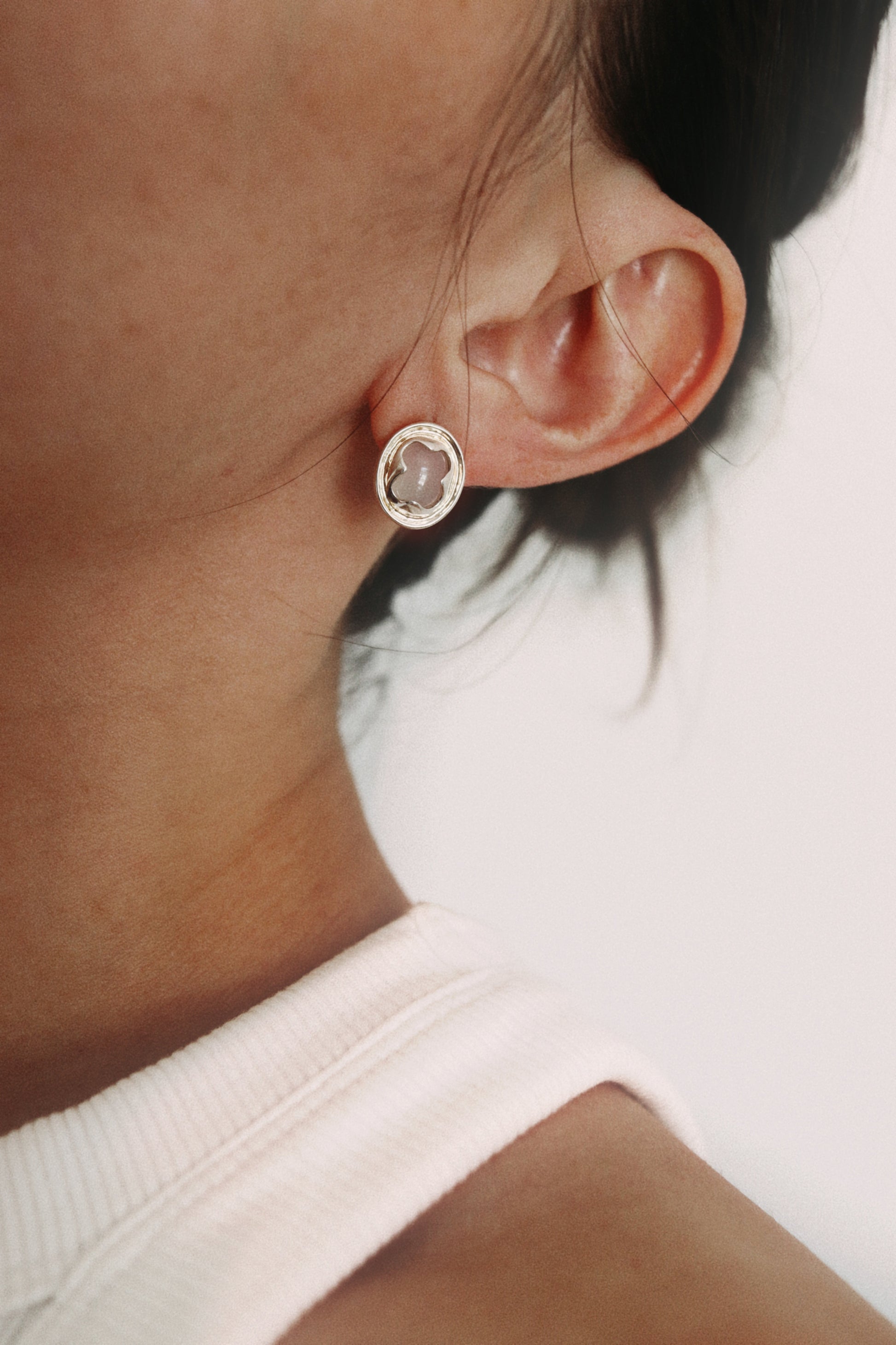 A close-up shot of a person's ear wearing a small oval stud earring with a silver-toned bezel and a pale pink stone.