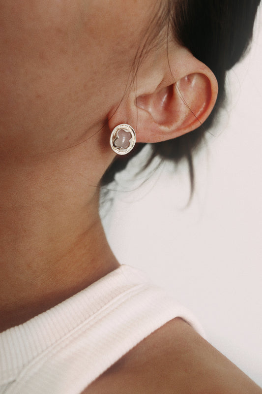 A close-up shot of a person's ear wearing a small oval stud earring with a silver-toned bezel and a pale pink stone.