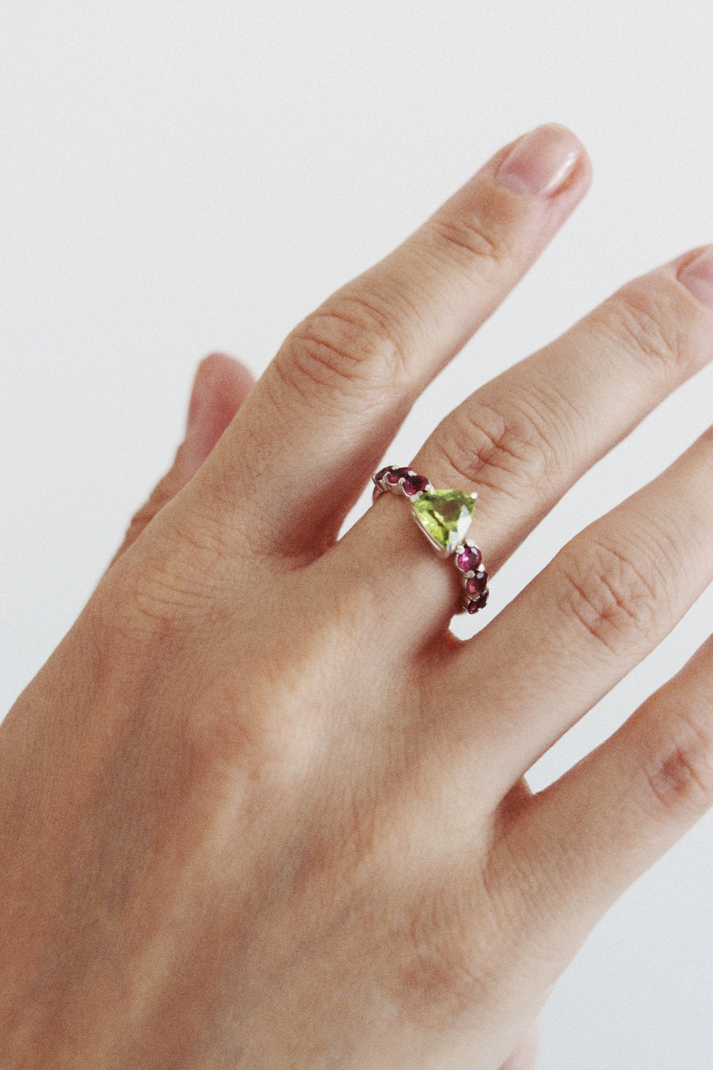A close-up of a hand wearing a twisted metal ring with a green pear-cut center stone and small pink gemstones along the band, set against a plain light background.
